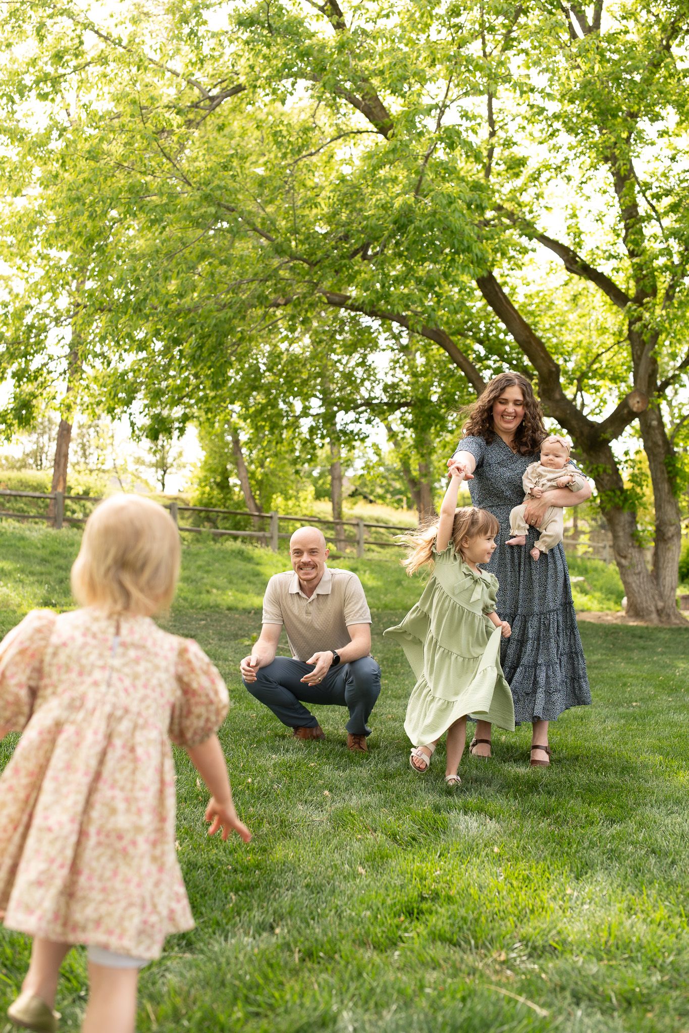 Happy family playing outdoors on green grass, parents watching children run toward them under lush trees on a sunny day.