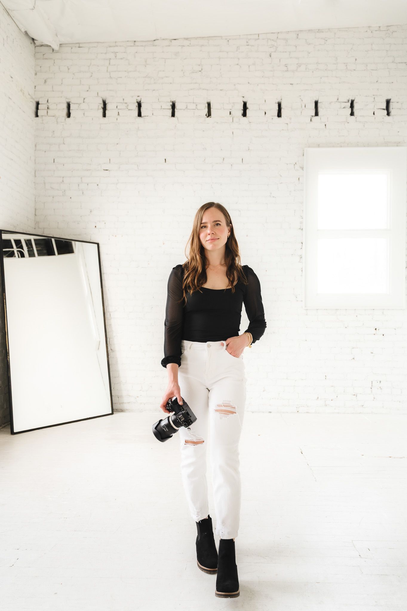 Fashion-forward photographer in black top and distressed white jeans holding camera in studio.