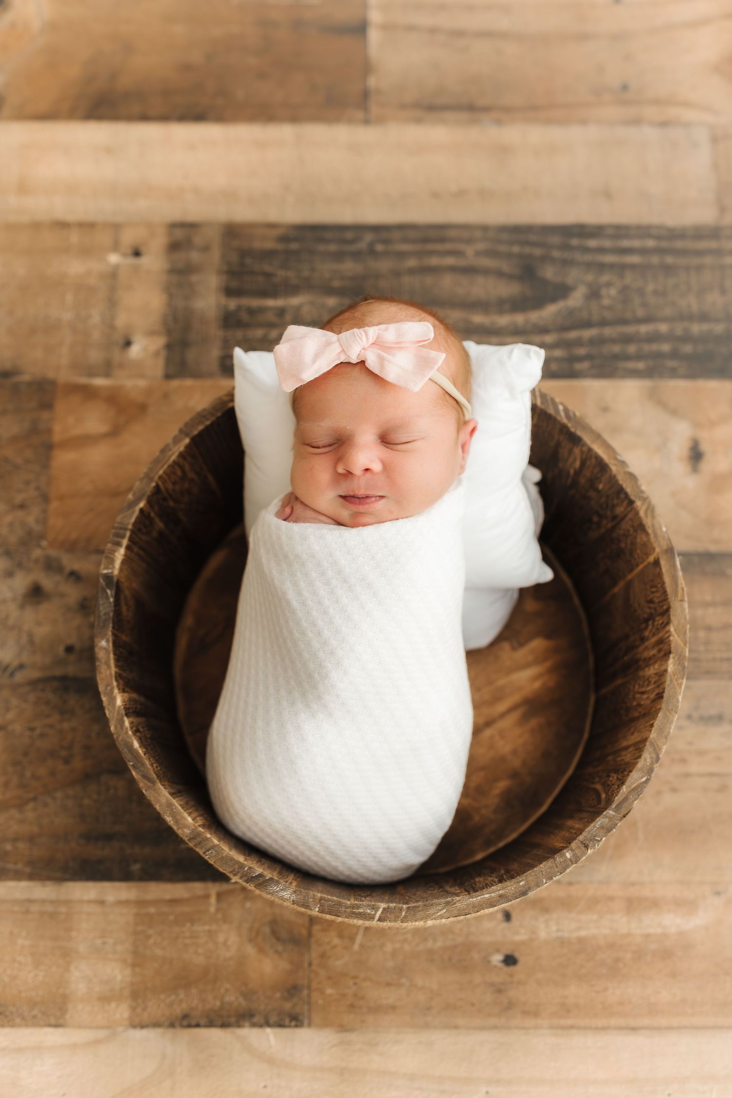 Newborn baby wrapped in white swaddle sleeping peacefully in wooden bowl.