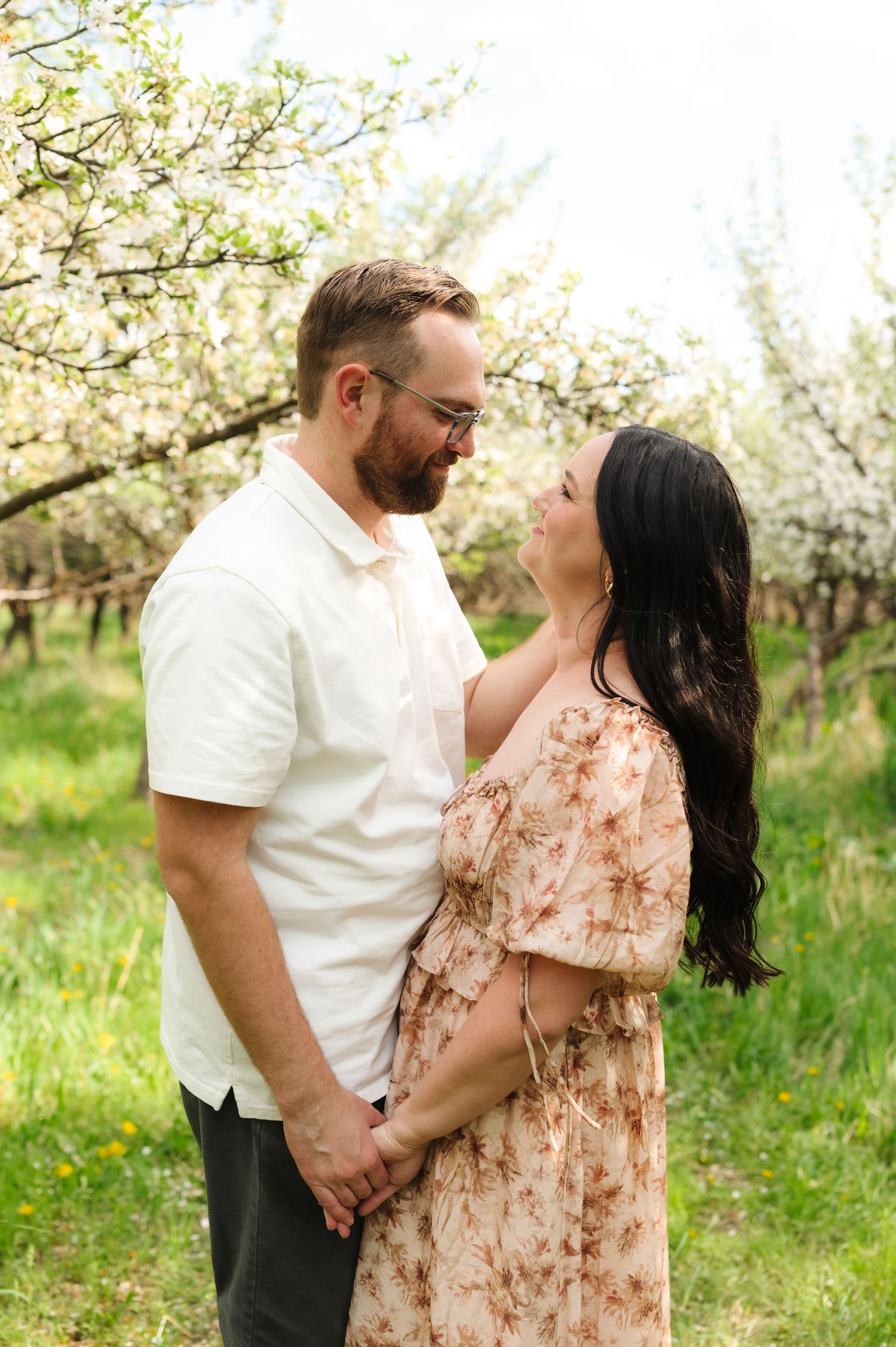 Romantic couple embraces in a blooming spring orchard with white blossoms and lush green grass.