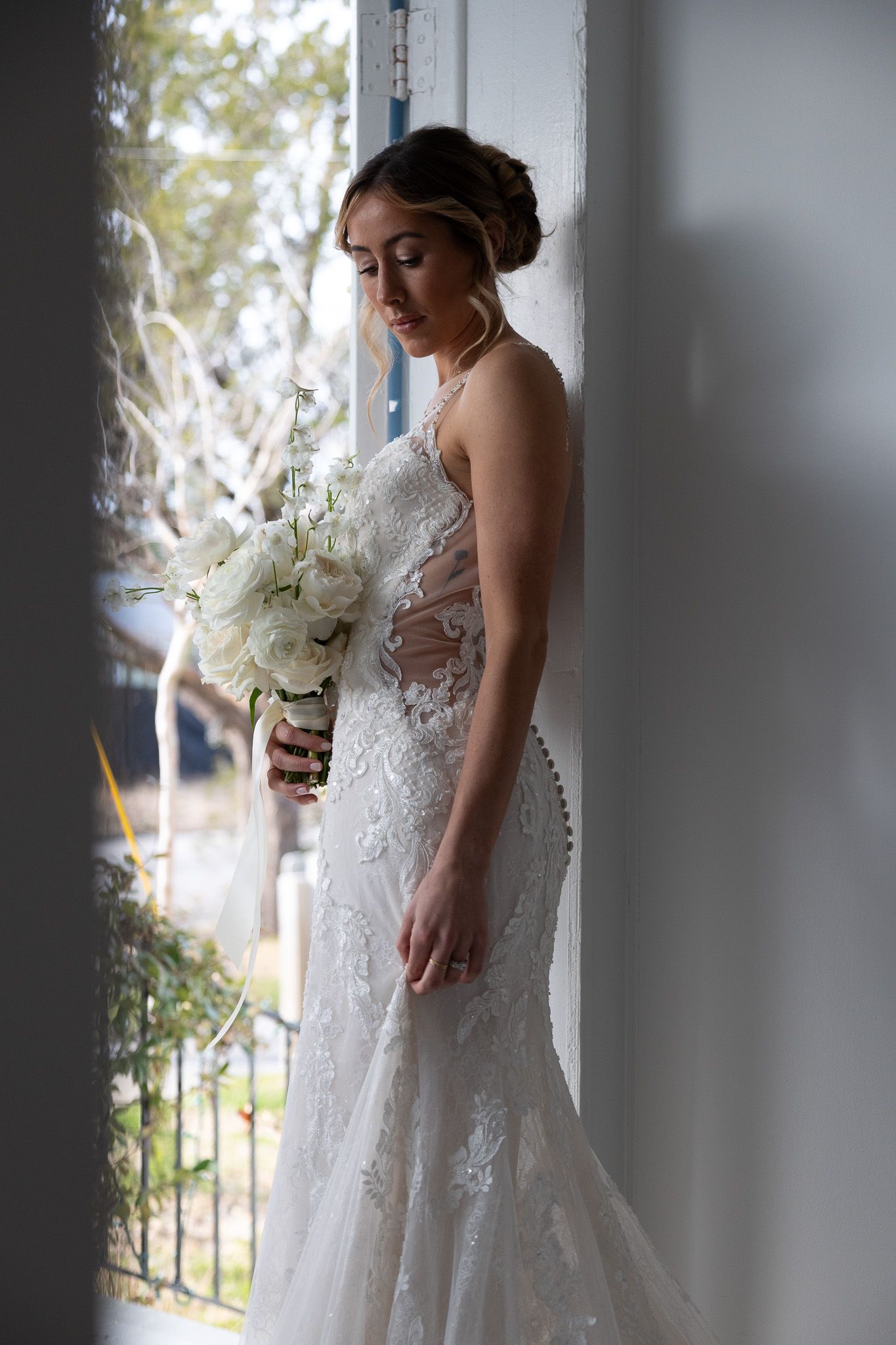 A bride in a lace gown stands pensively by a window holding a white bouquet.