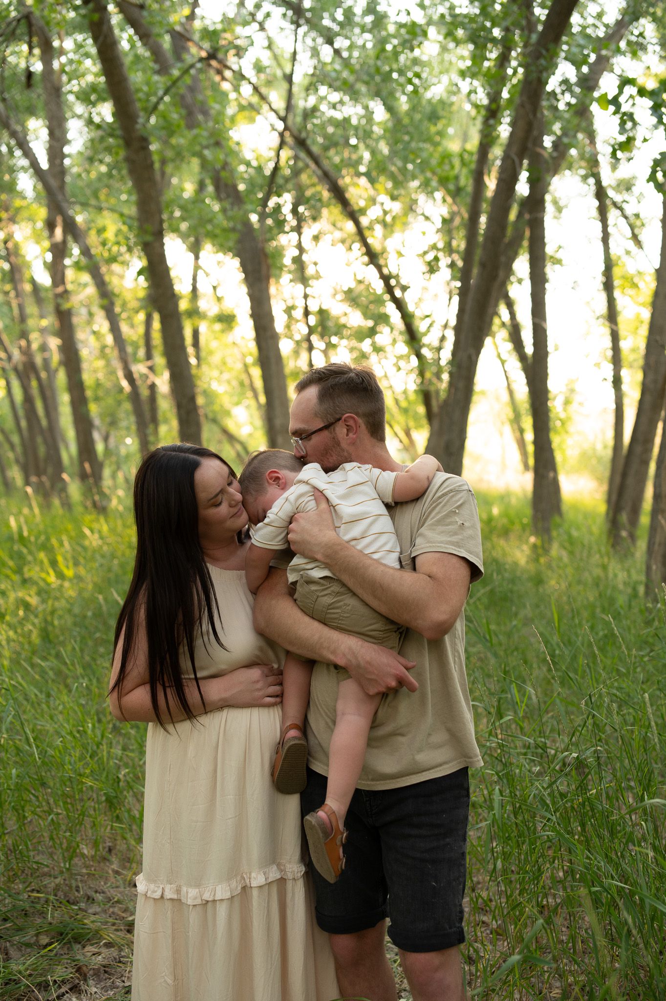 A young family with a newborn baby shares a tender moment on a lush green forest path during golden hour.