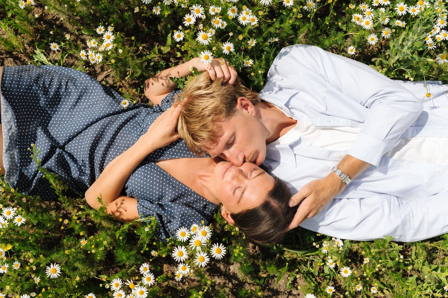 Romantic couple lying in a daisy field, sharing a tender kiss surrounded by white wildflowers on a sunny day.