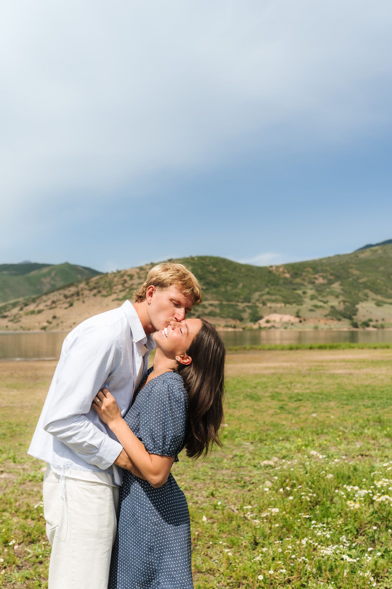 Couple embracing and kissing outdoors near a lake with green hills and blue sky in background.
