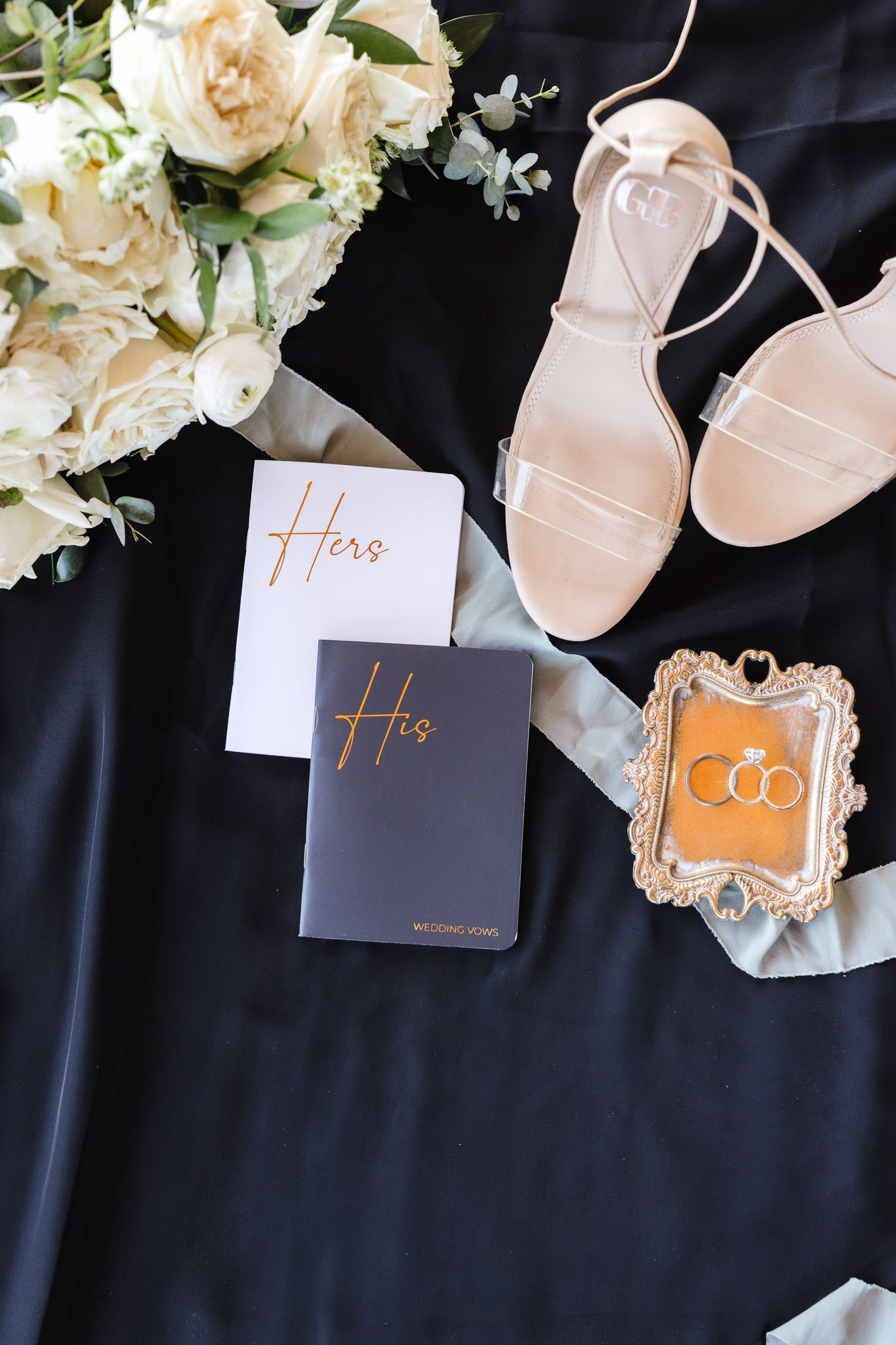 Wedding details featuring elegant pink and white floral arrangements with his and hers tags and delicate sandals on black fabric.