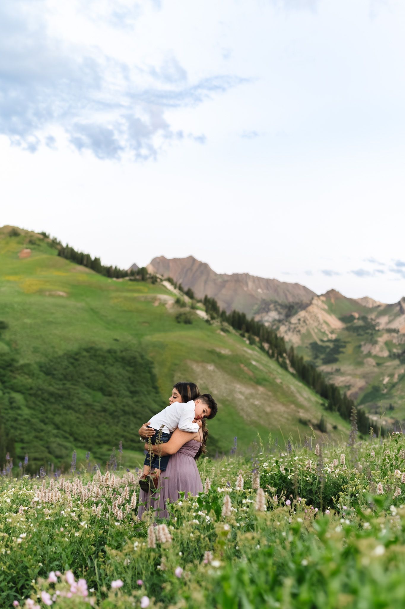 Mother holding child in wildflower meadow with green mountain slopes and cloudy sky backdrop.