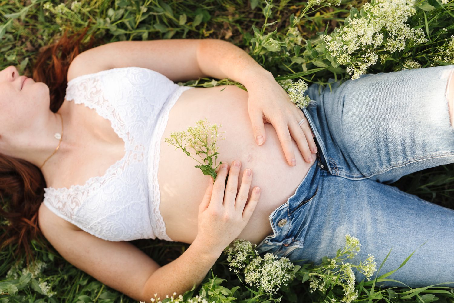 Pregnant woman lying in green grass wearing white lace bralette and jeans, holding wildflowers on her baby bump.