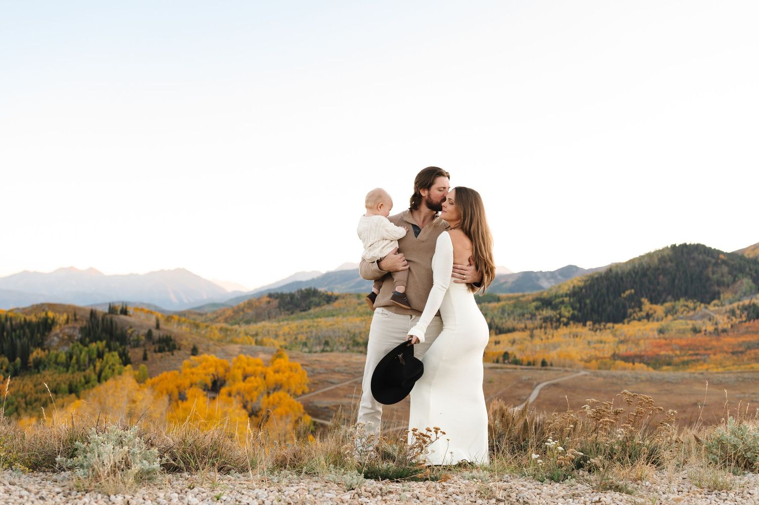 Couple kissing while holding baby on mountain overlook with colorful autumn foliage and peaks in background.