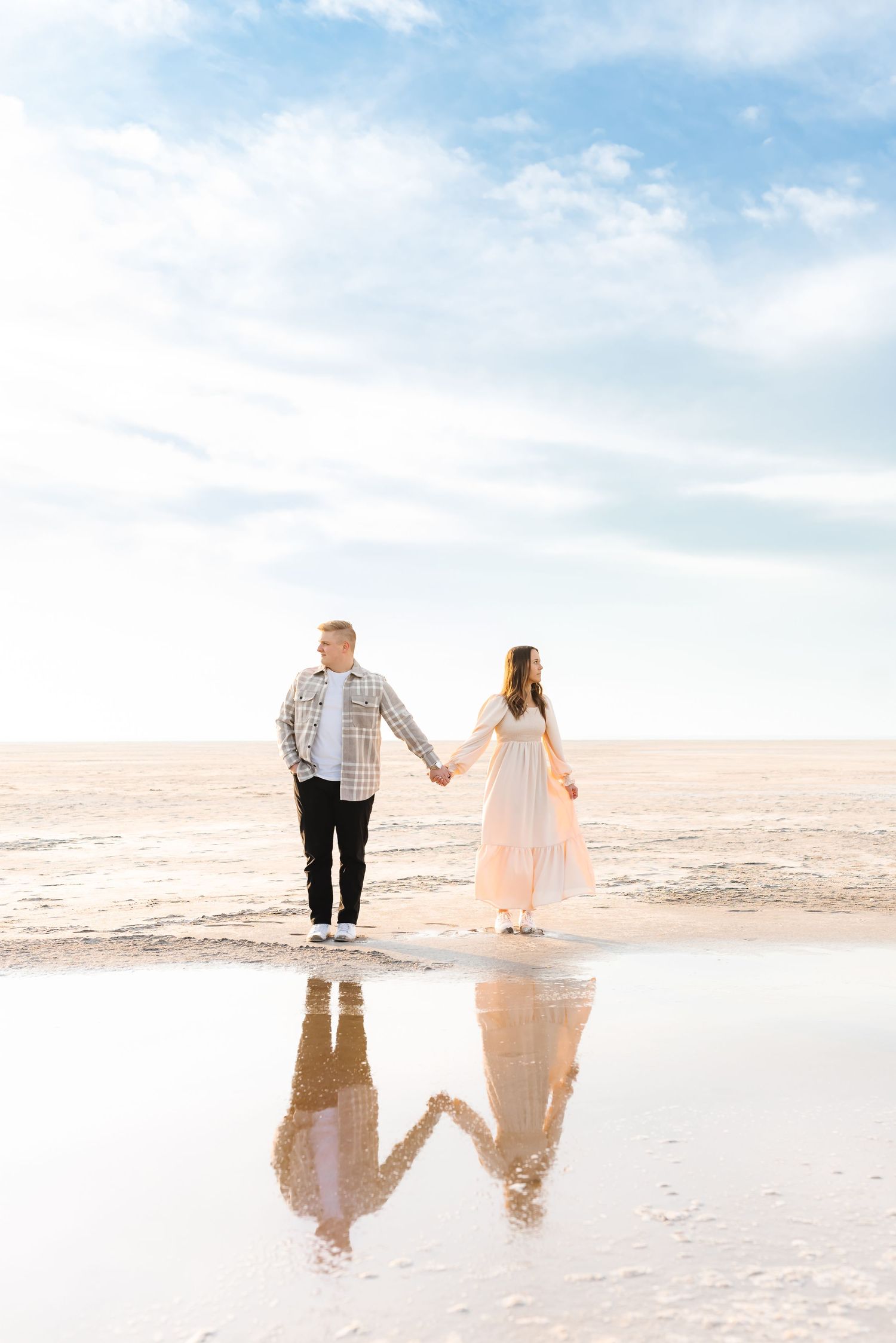 Couple holding hands on salt flats with reflection in water during sunset.