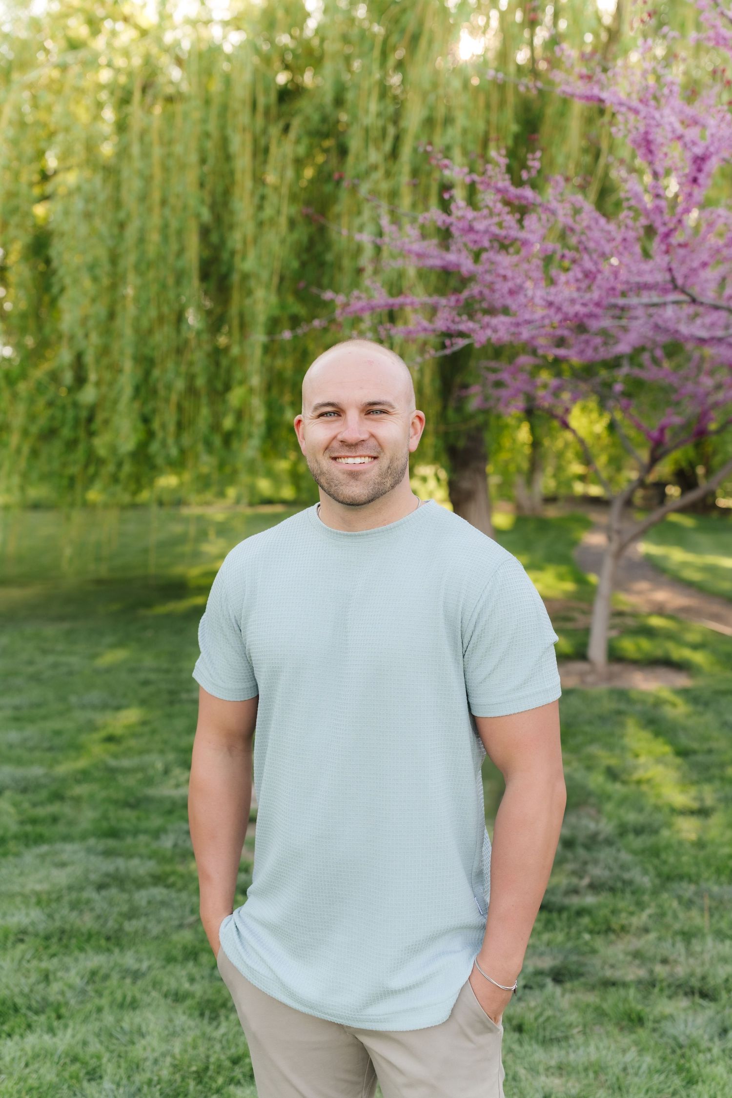 A person wearing a light blue t-shirt stands outdoors with blooming pink cherry blossom trees and green grass in the background.
