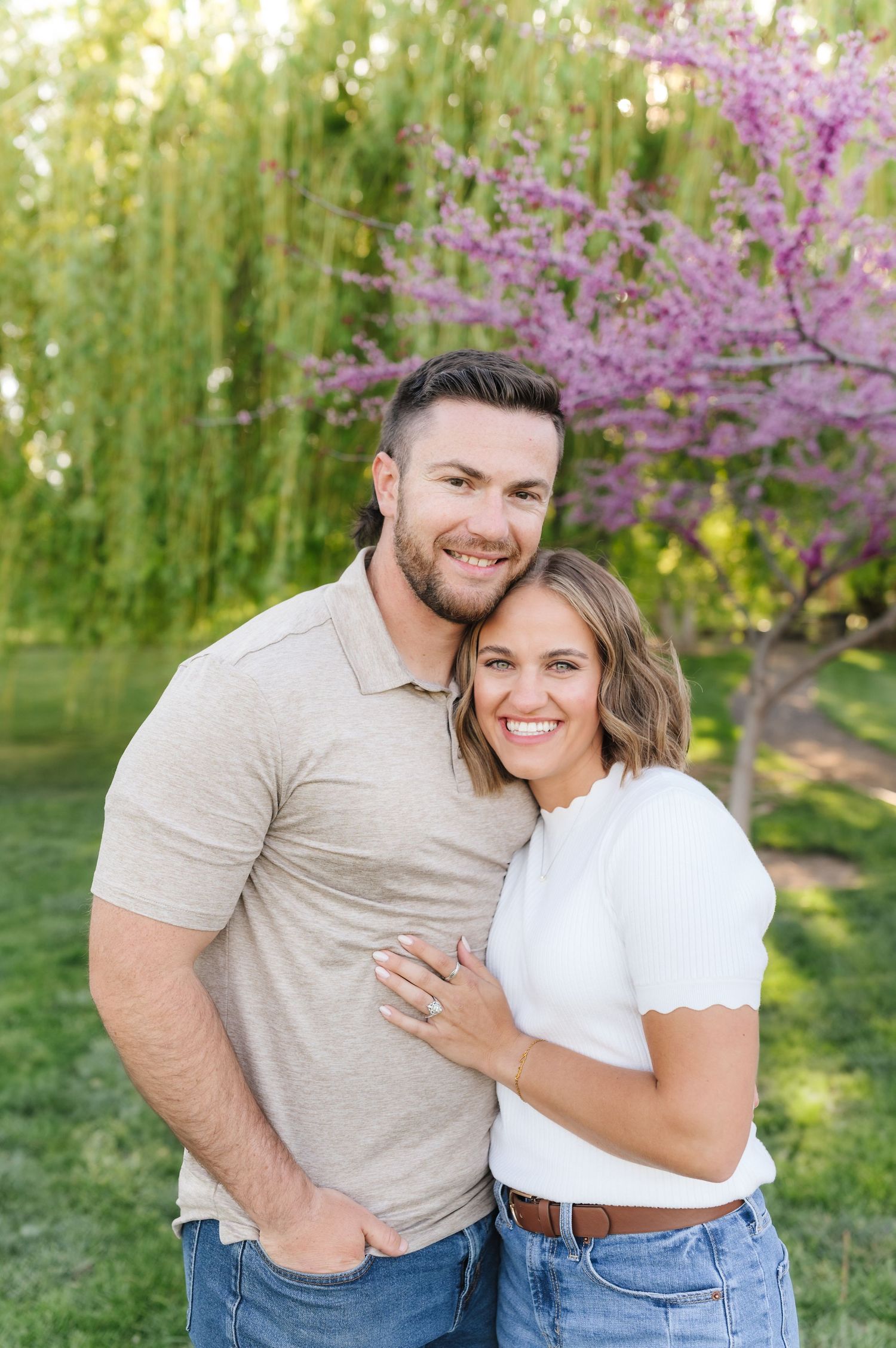 Couple embracing outdoors with purple flowering trees and willow branches in background.