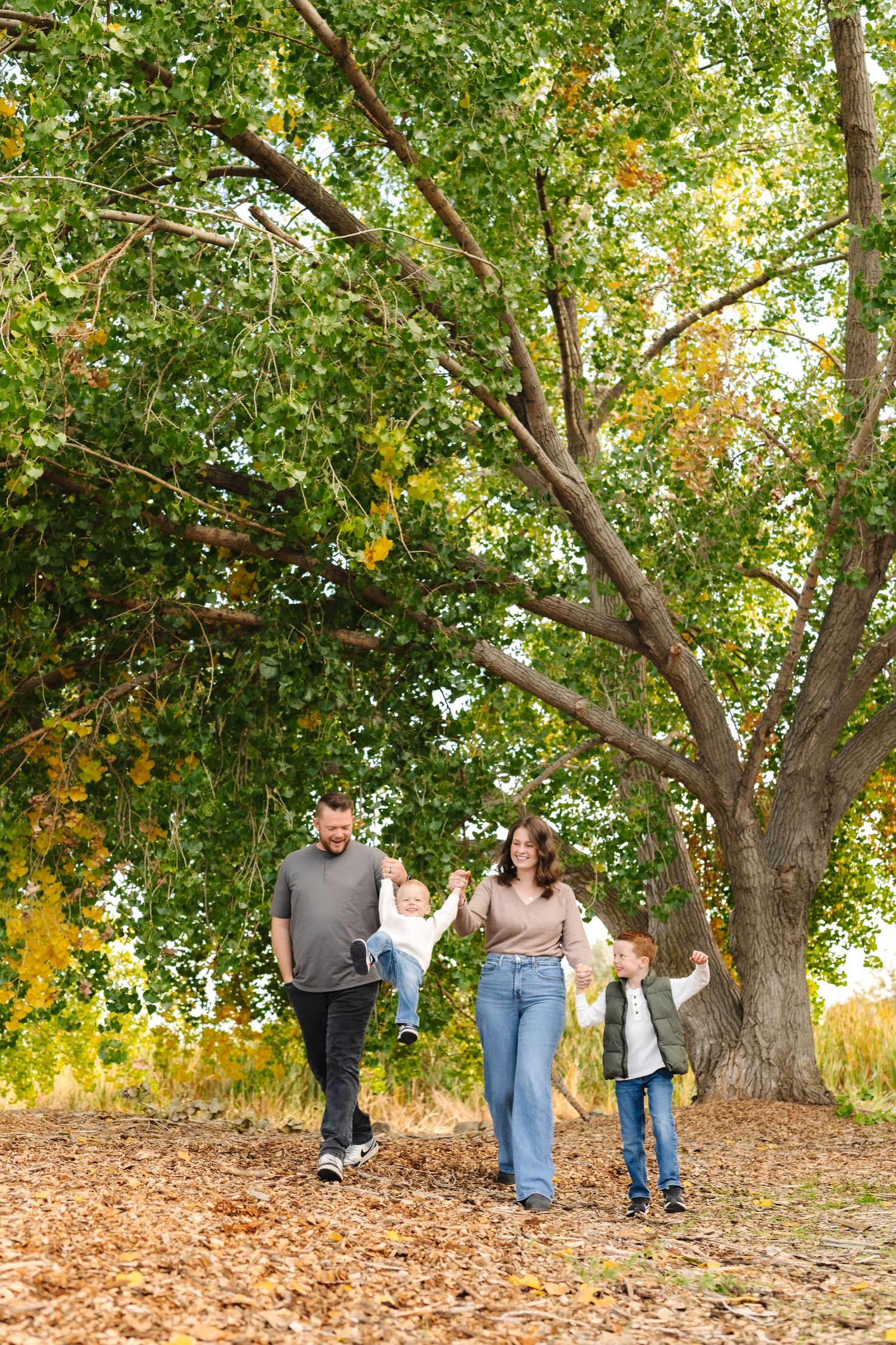 Family walking among fallen autumn leaves under large green trees, parents swinging young children by their hands.