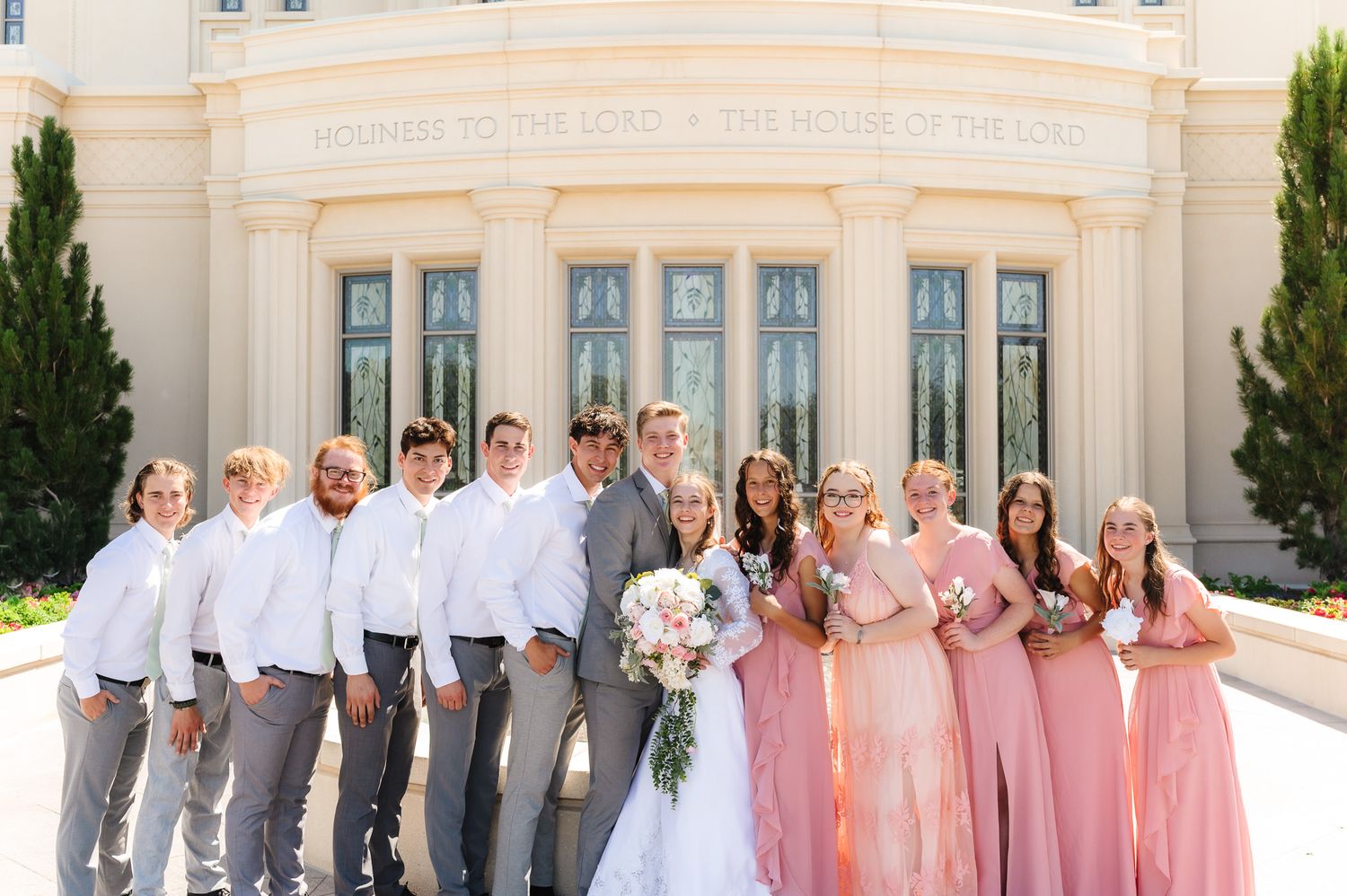 Wedding party poses outside a temple, bride in white, bridesmaids in pink gowns, groomsmen in gray suits.