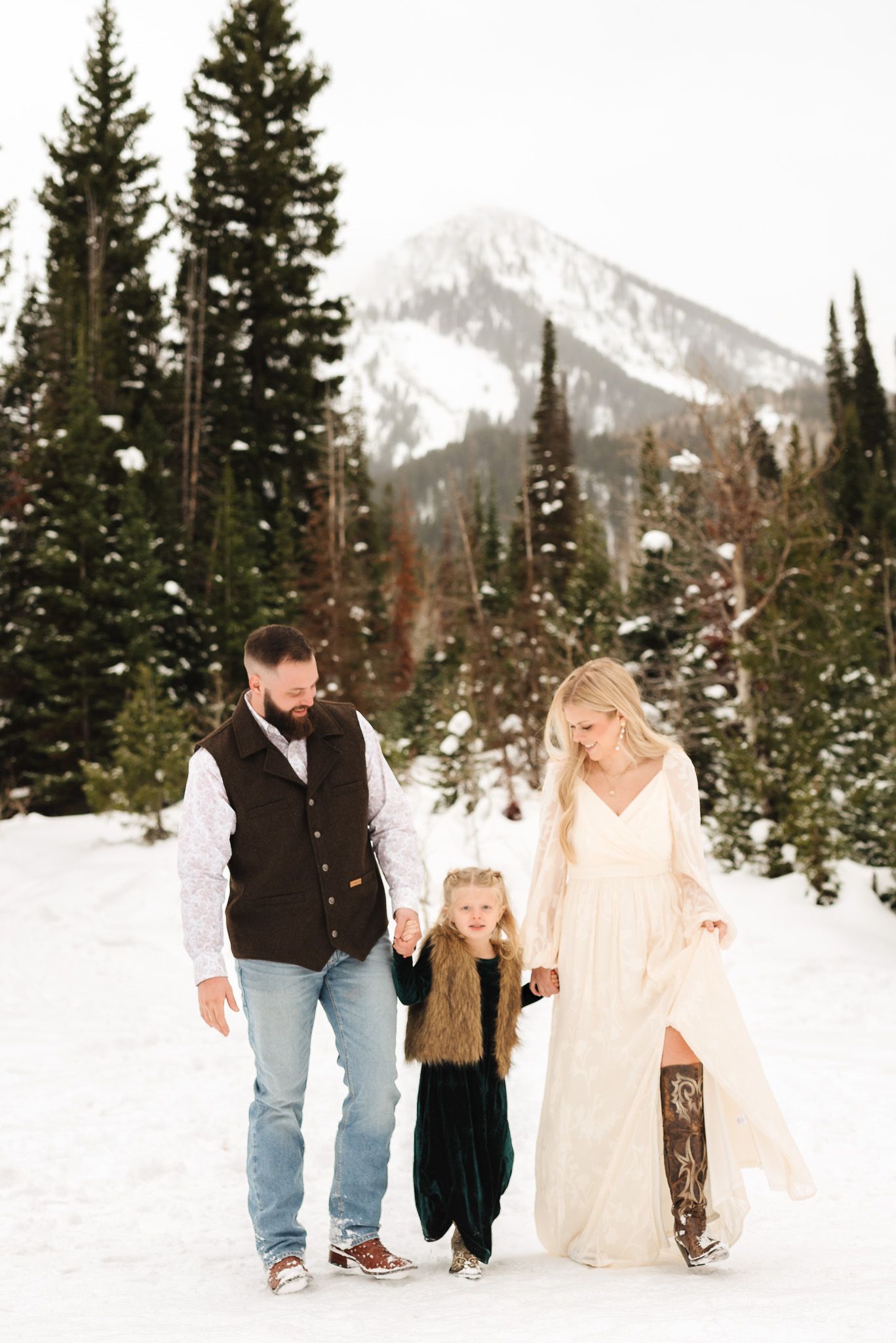 Family of three walks hand-in-hand in snowy mountain landscape, bride in white gown, groom in vest, child in fur coat.