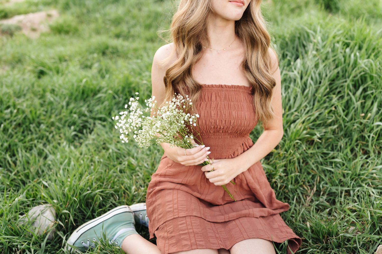Young woman in rust-colored smocked dress sitting in green grass holding a bouquet of white wildflowers.