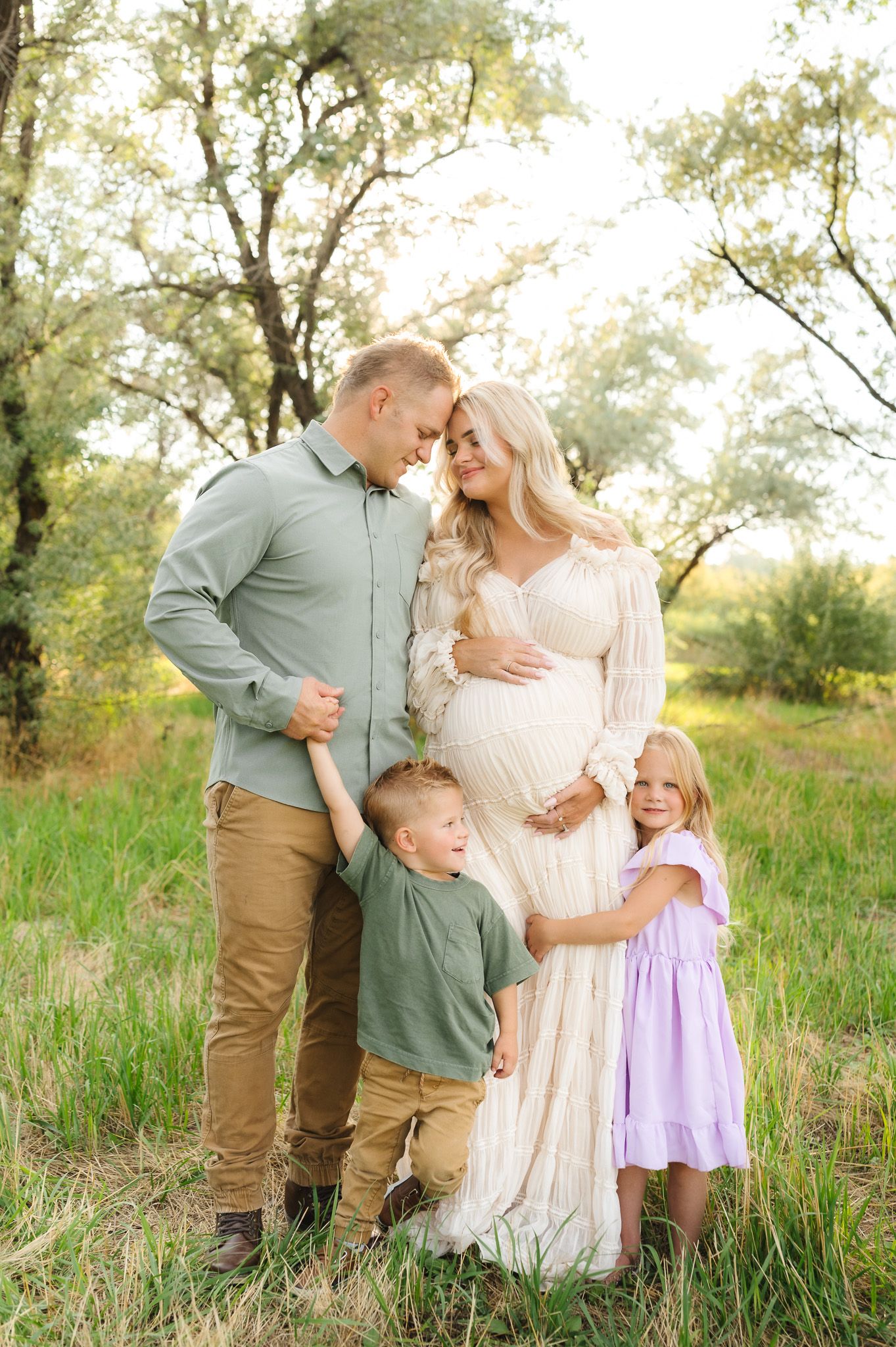 Family portrait session in a sunlit meadow with tall grass and trees in the background.