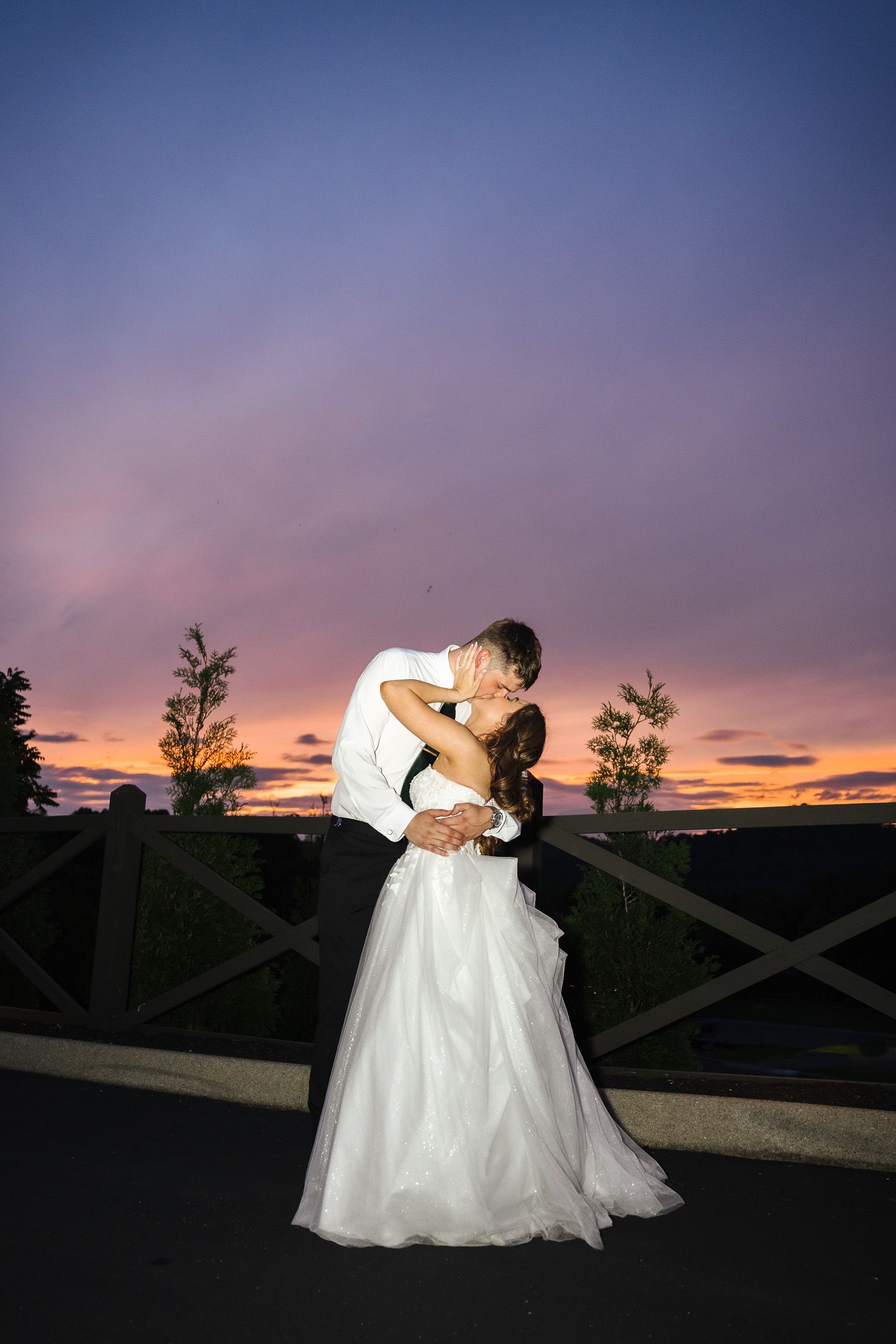 Bride and groom sharing a romantic kiss at sunset, with vibrant purple and orange sky backdrop at outdoor wedding venue.