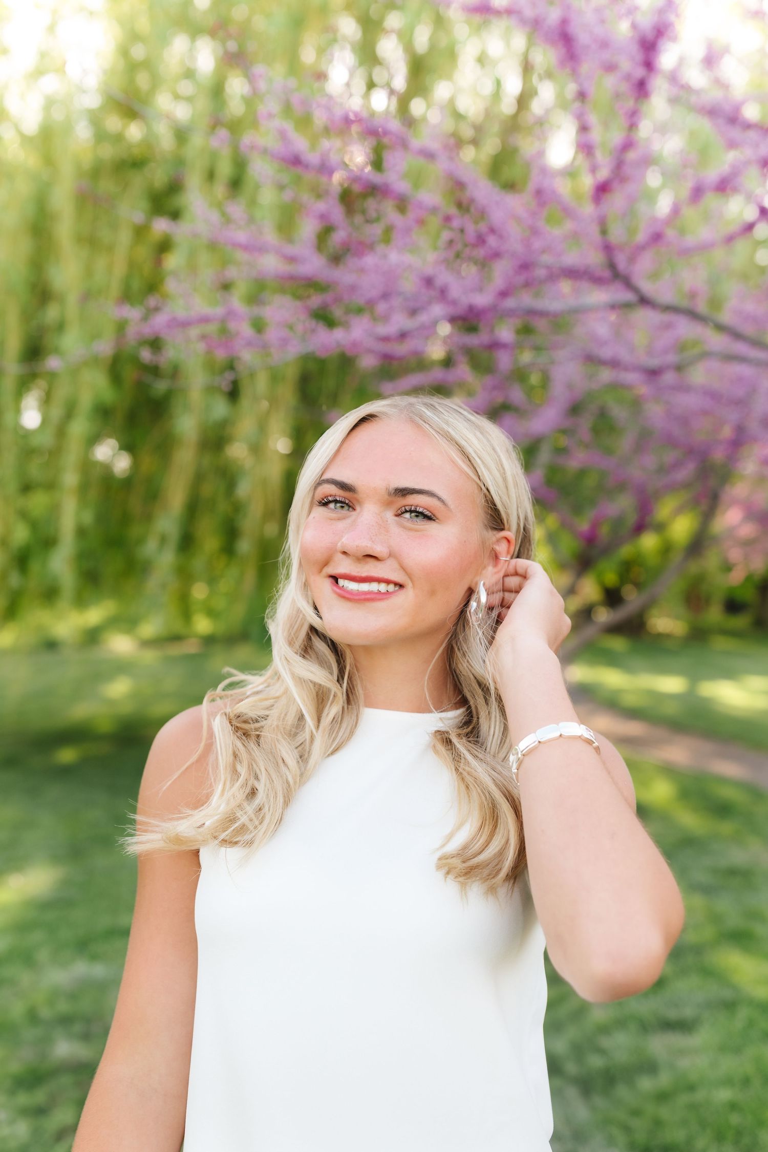 Woman in white top smiles radiantly against backdrop of spring blossoms and willow trees.