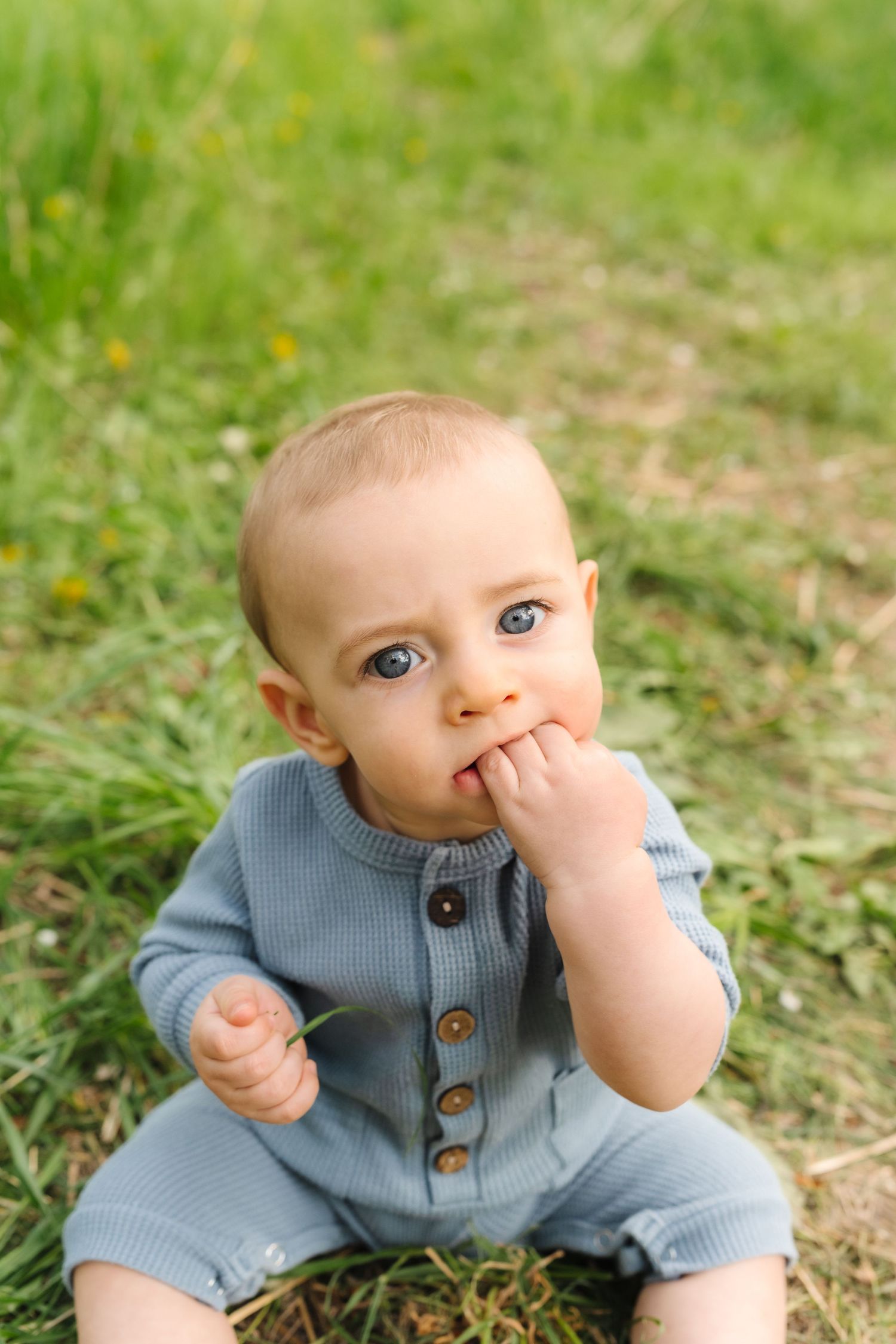 Baby wearing blue knit sweater sits in grass while making playful expressions.