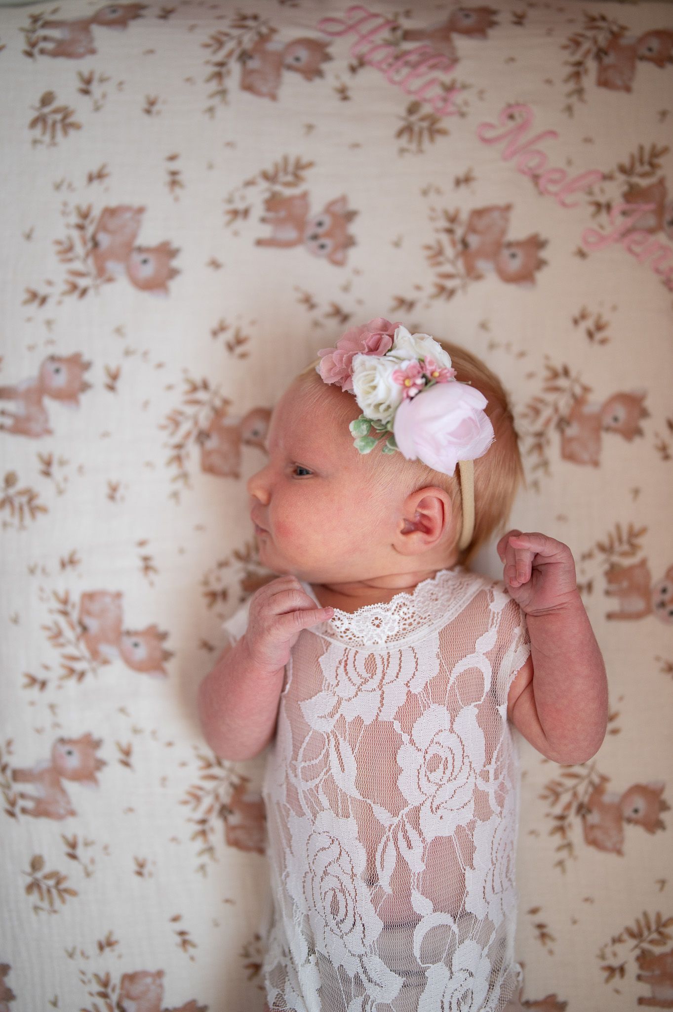 Newborn baby girl in white lace outfit with pink floral headband lying on deer print blanket.