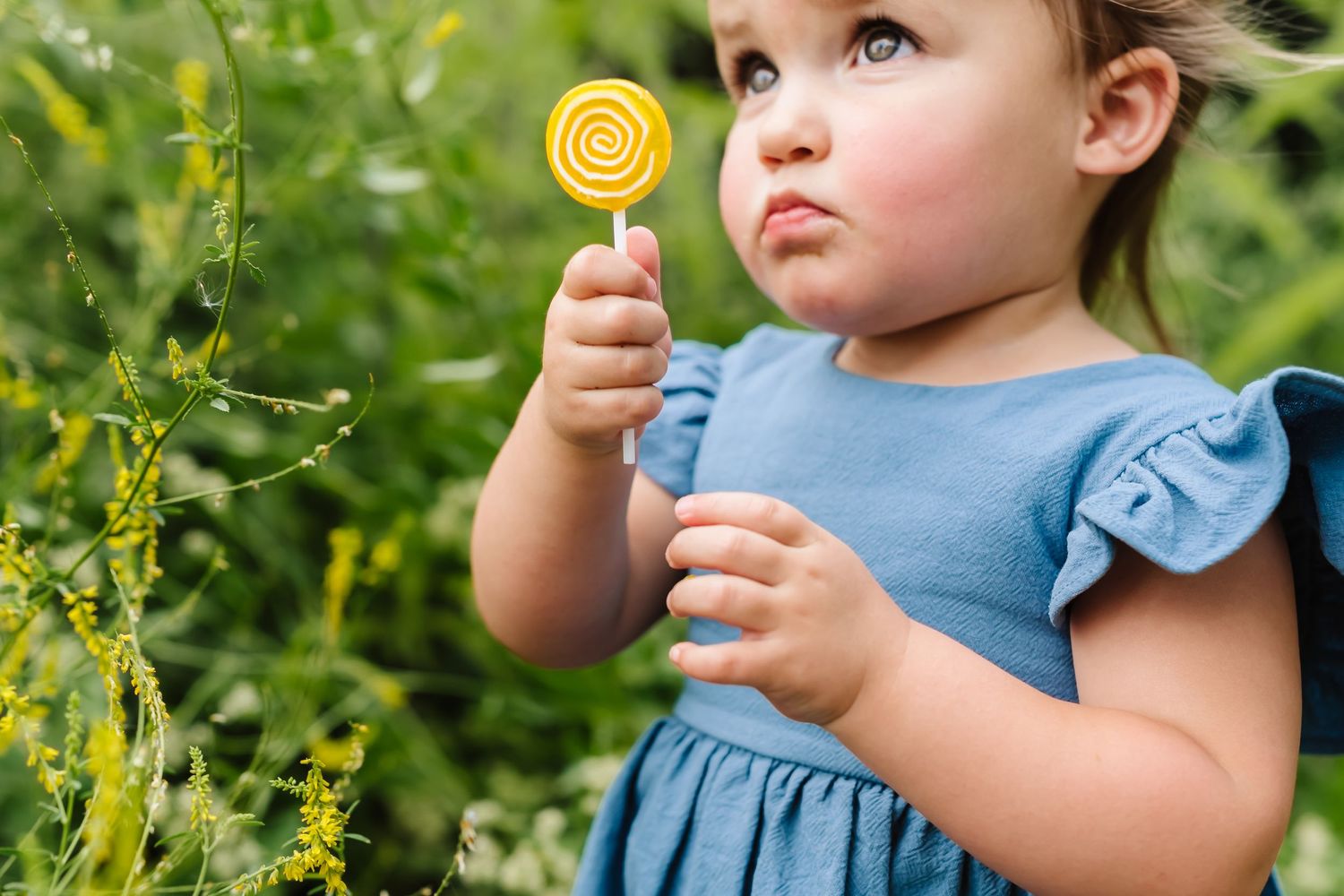Small child in a blue dress holding a yellow lollipop while standing in a garden.