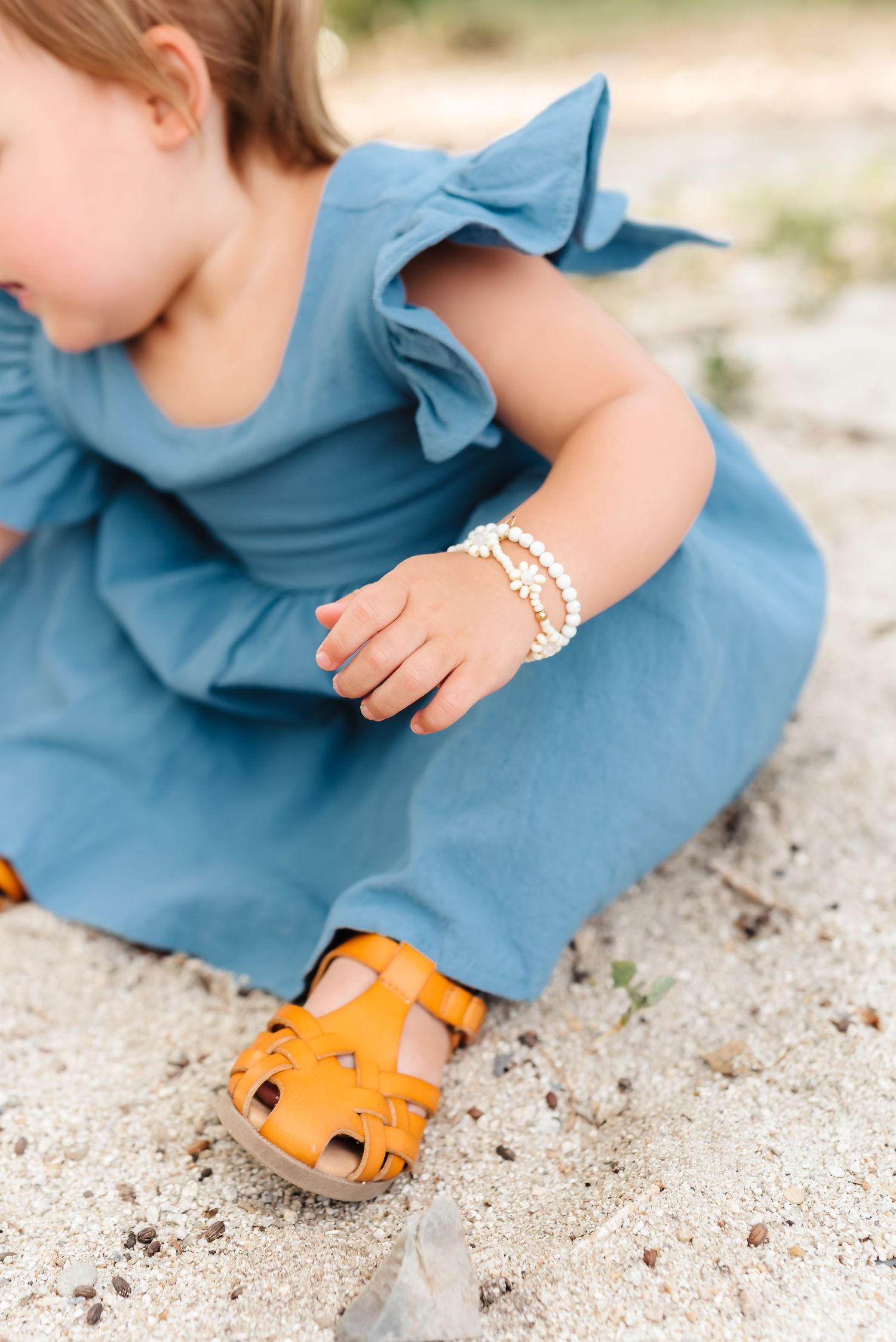 Close-up of small orange sandals on sandy ground with blue dress fabric visible.