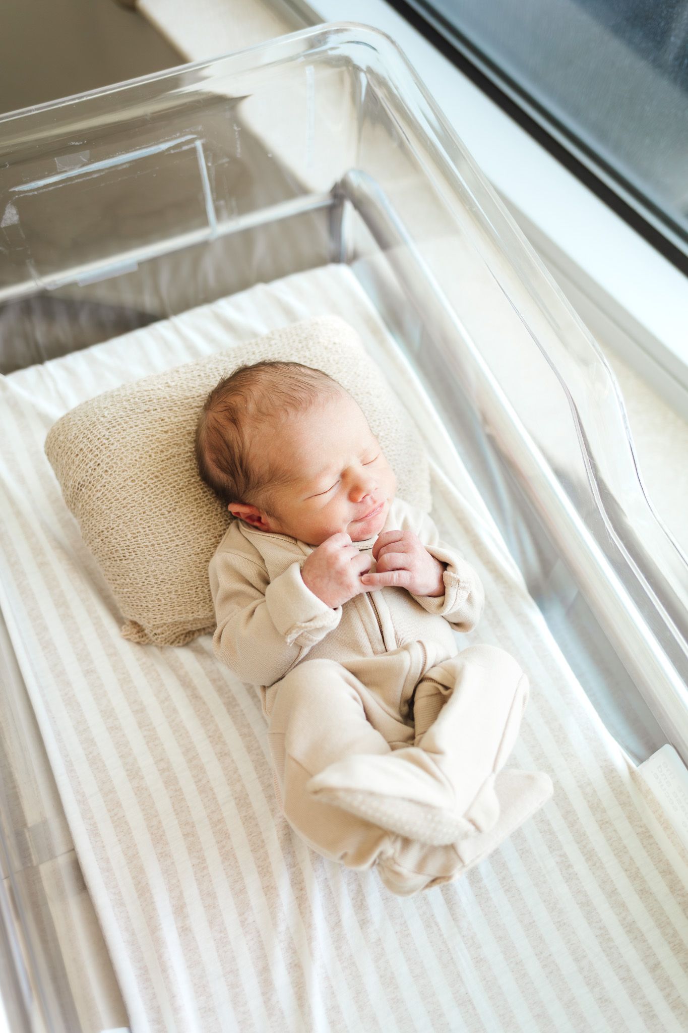 Newborn baby sleeping peacefully in a hospital bassinet, dressed in a beige onesie on a striped white sheet.