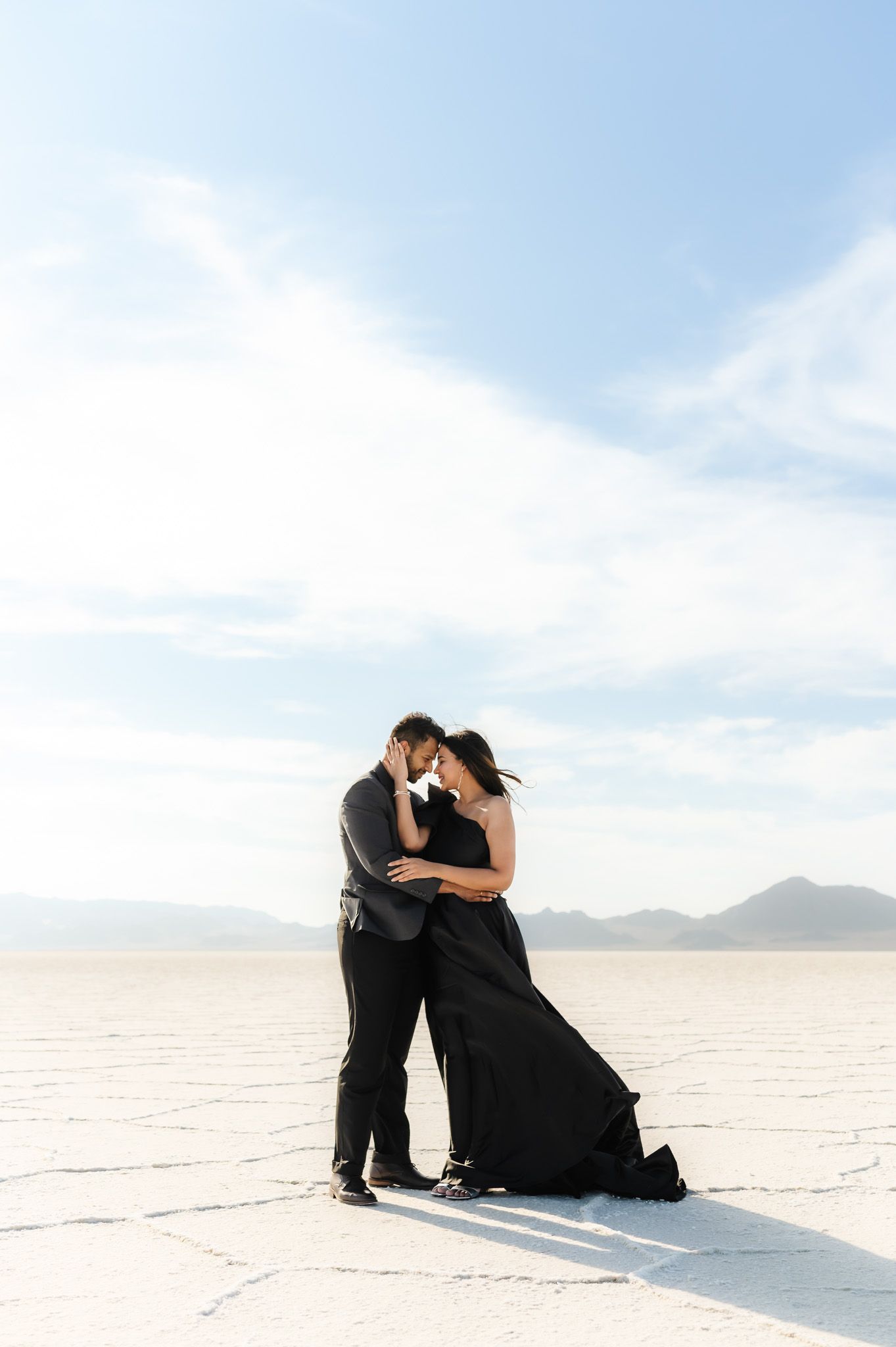Romantic couple in black outfits embracing on a snowy white landscape with mountains in the background.
