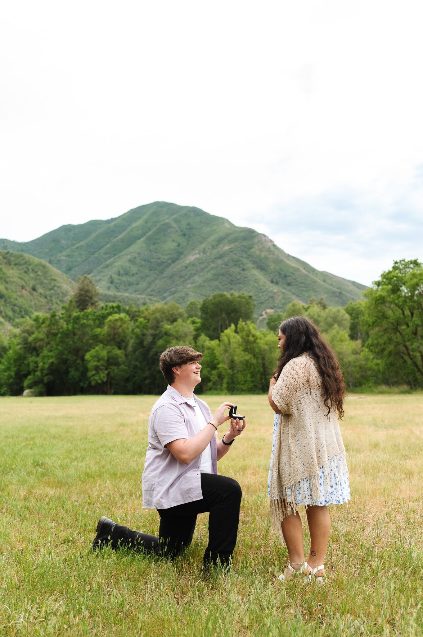 Man kneeling on one knee proposing to woman in a green mountain meadow with ring box.