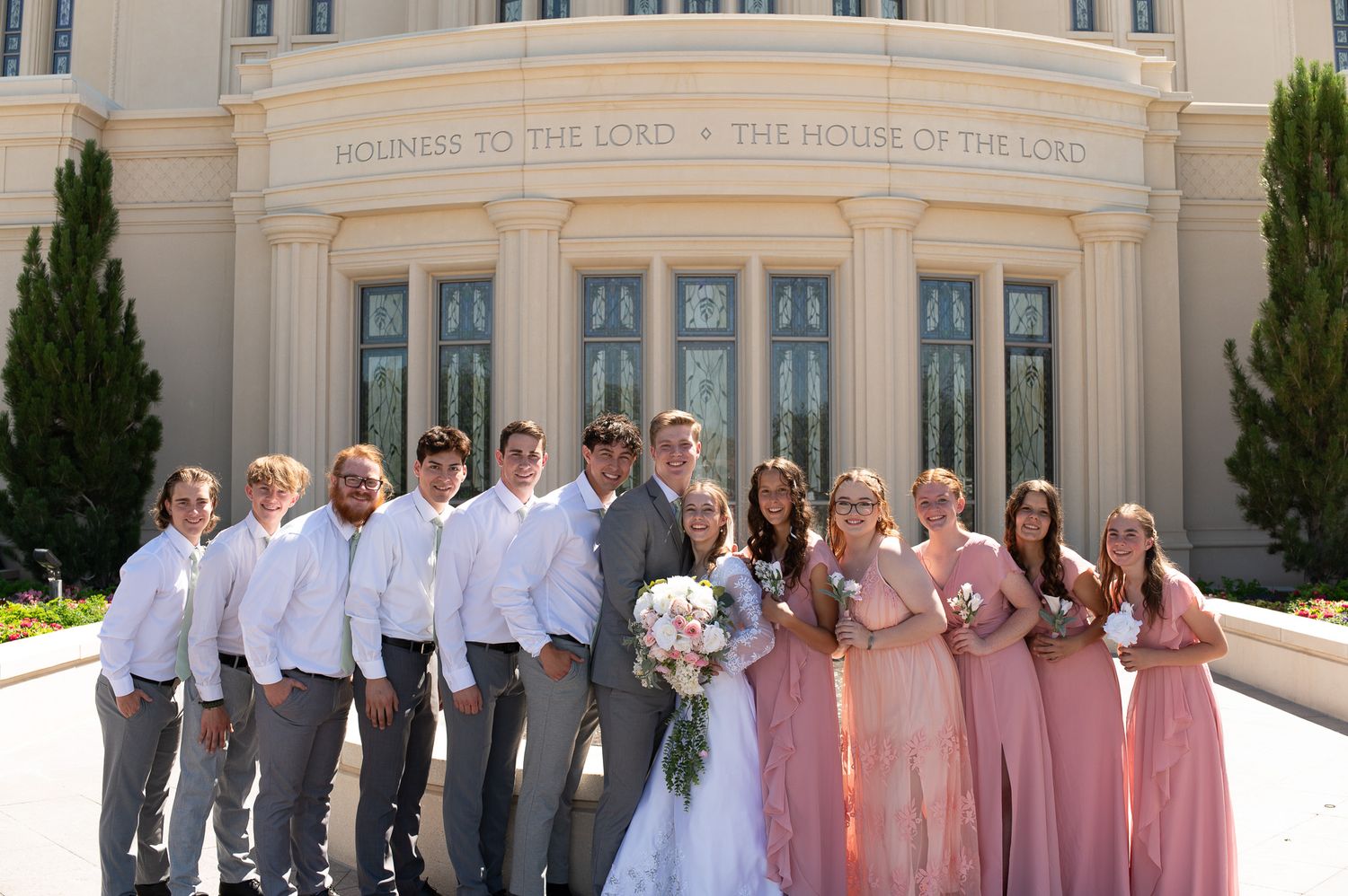 Wedding party posing outside an LDS temple, bridesmaids in pink gowns and groomsmen in gray suits with the bride and groom.