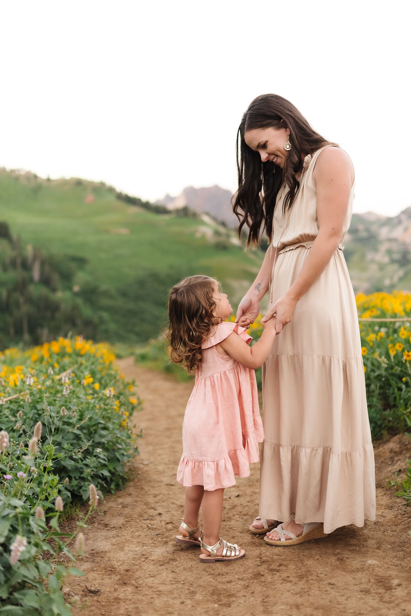 A mother in a cream dress holds hands with her toddler daughter among yellow wildflowers on a mountain path.