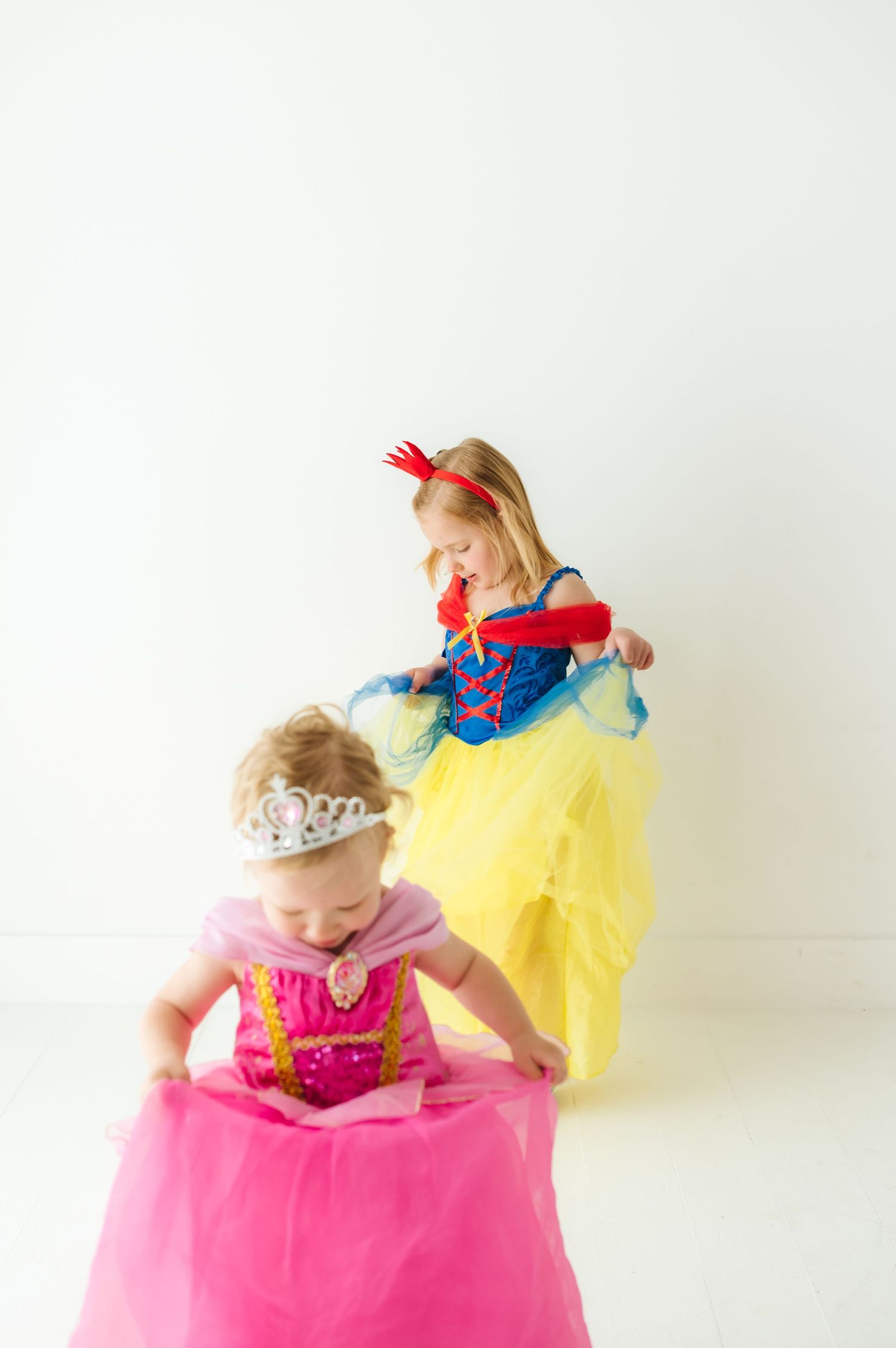 Two children in princess costumes, one in yellow and one in pink, playing together against white background.
