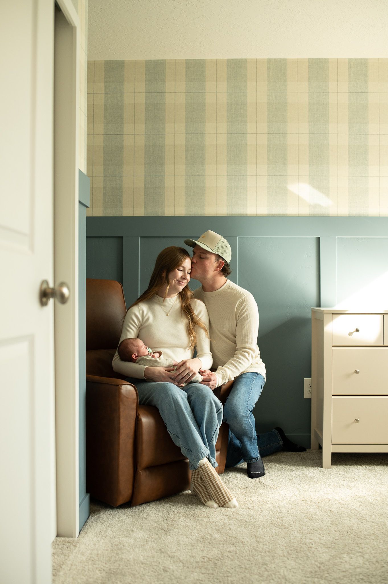 A couple shares a tender moment in a nursery, sitting together on a leather chair holding their newborn baby.