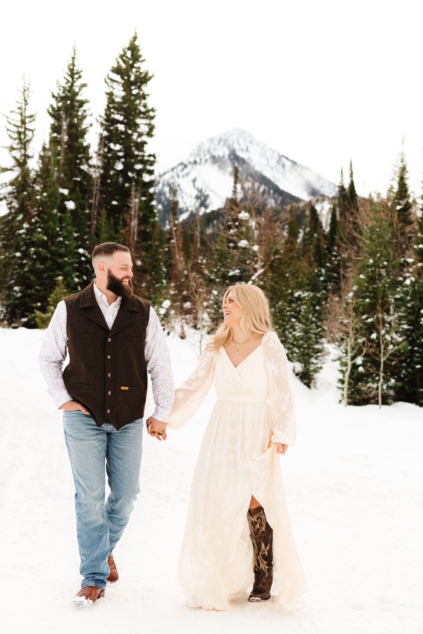 Couple walking hand in hand through pristine snow with evergreen forest backdrop.