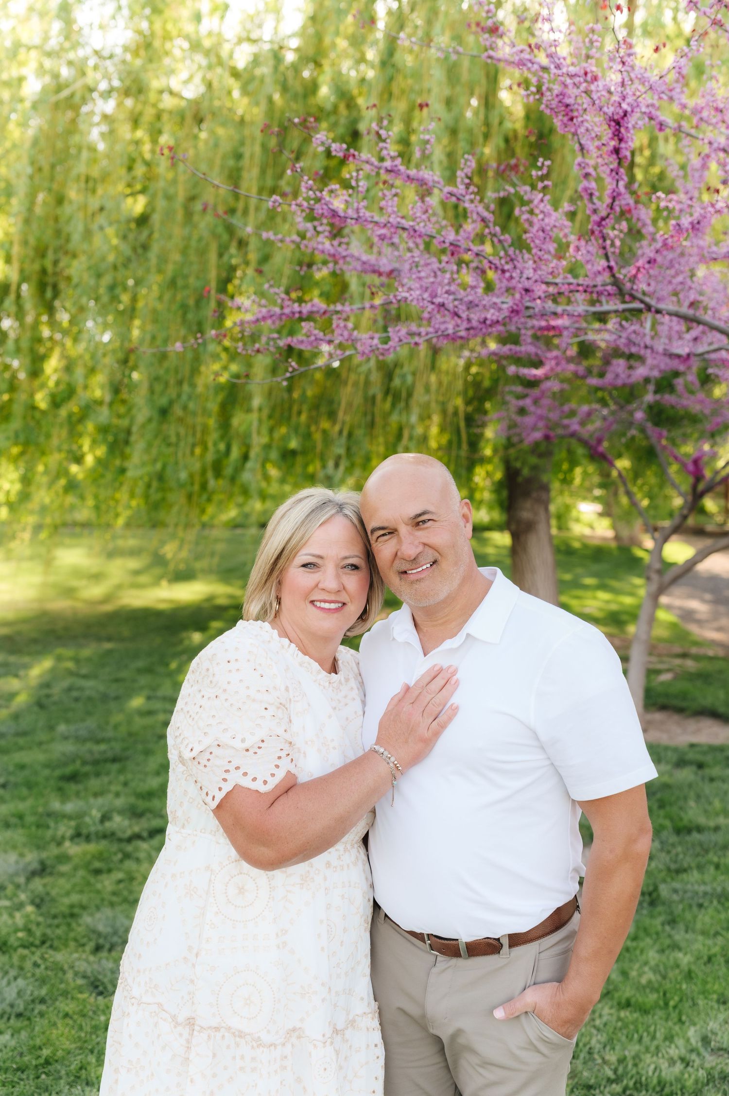 Couple in white and beige outfits embracing under flowering cherry blossom tree.