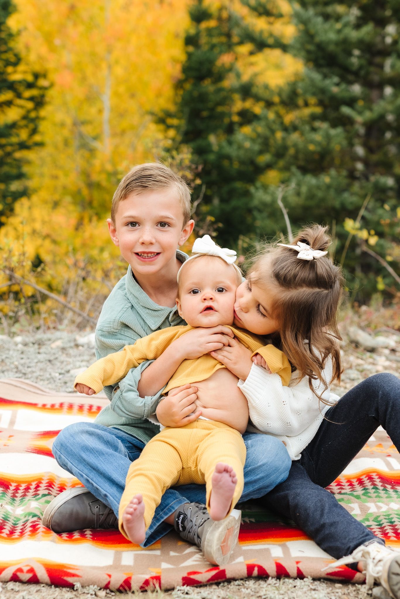 Siblings share sweet moments on striped blanket with golden autumn trees in background.