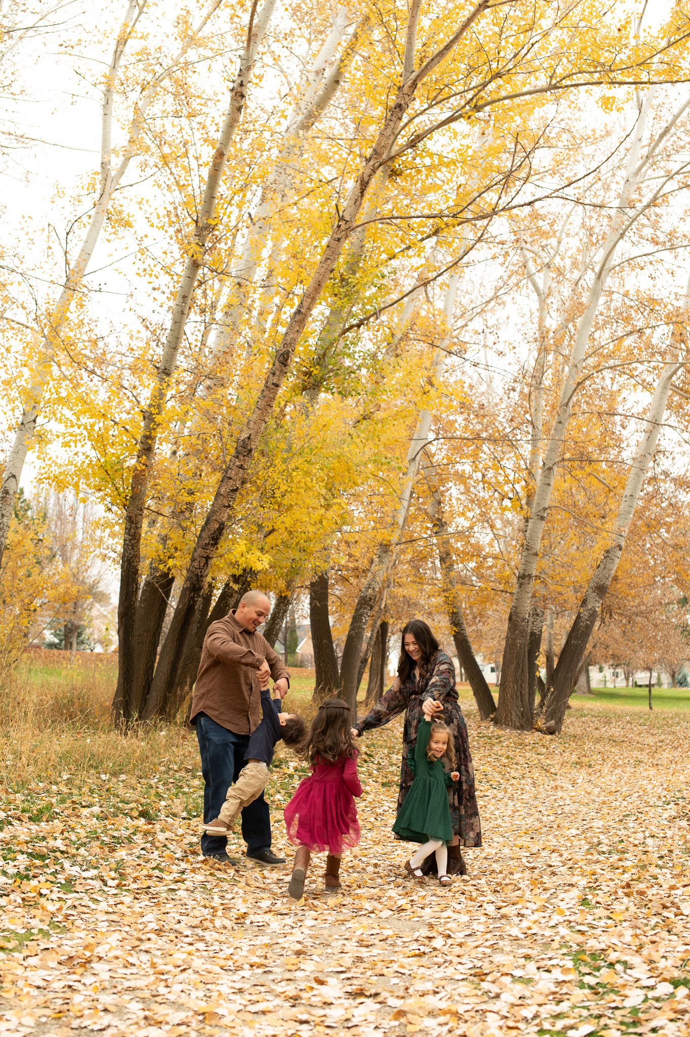 Family playing together on a leaf-covered path surrounded by golden autumn trees in a park.