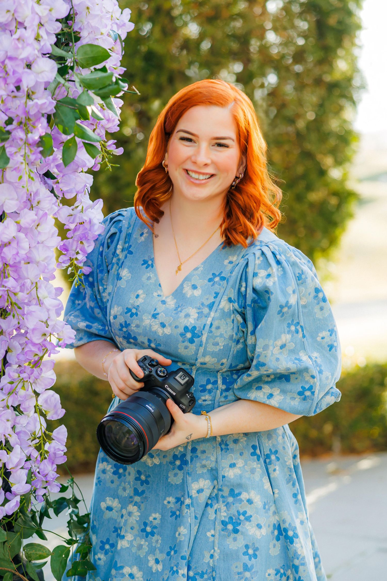 A photographer in a blue floral dress stands beside blooming purple wisteria while holding a professional camera.