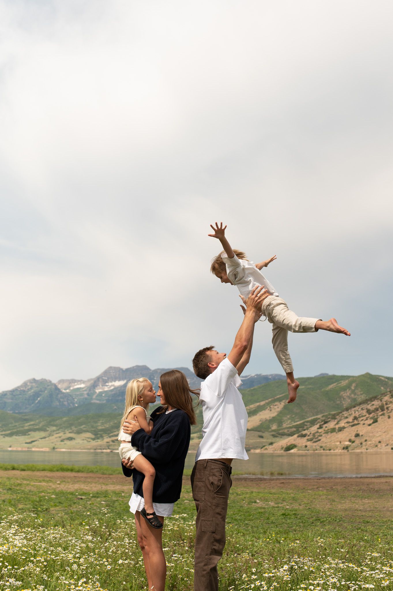 Family playing outdoors near a mountain lake, father lifting child in the air while mother holds another child.