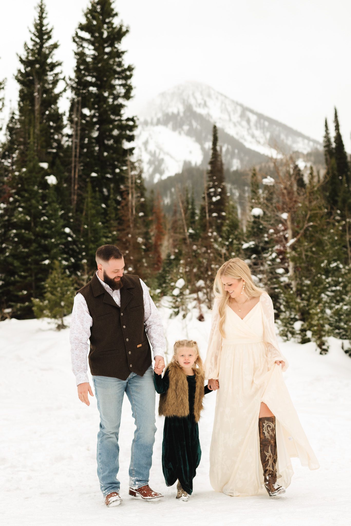Family of three walks hand-in-hand in snowy mountain landscape, woman in elegant white gown, man in vest, child in velvet dress.