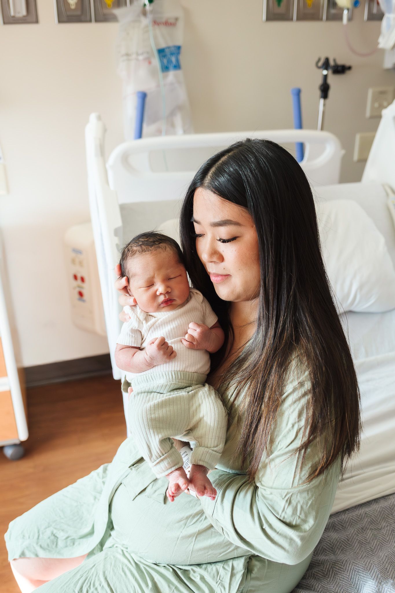 New mother in green pajamas tenderly holds her sleeping newborn baby in a hospital room after delivery.