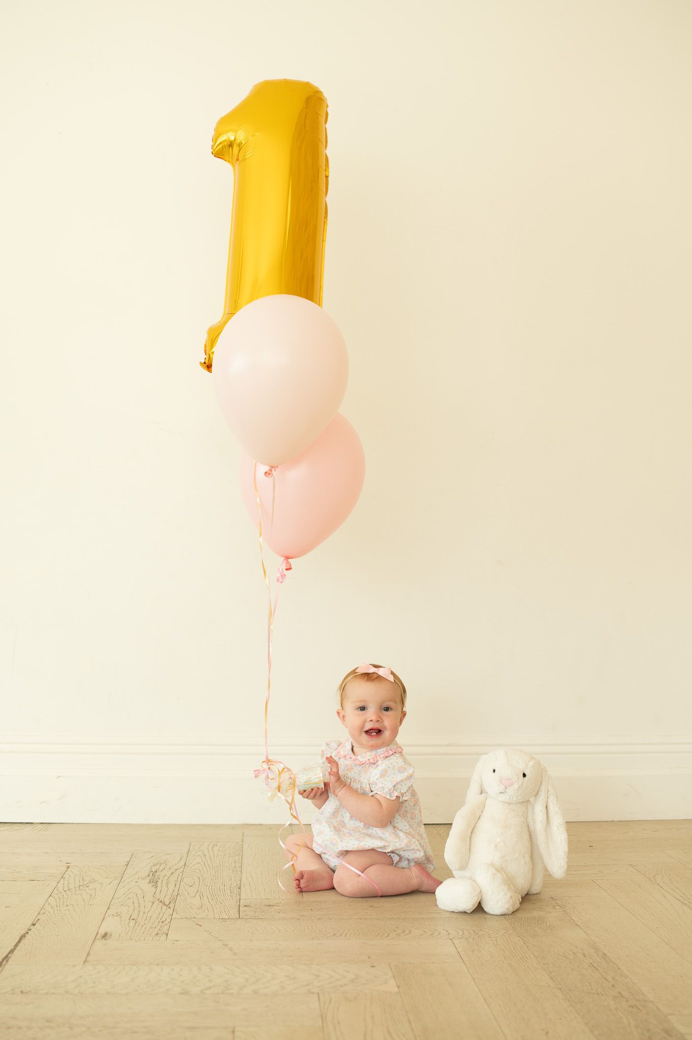 Baby girl in white dress holds pink and gold number one balloons with stuffed bunny for first birthday photo.