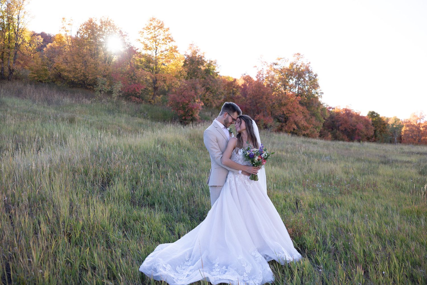 Wedding couple share a romantic moment in a golden meadow surrounded by colorful fall trees at sunset.