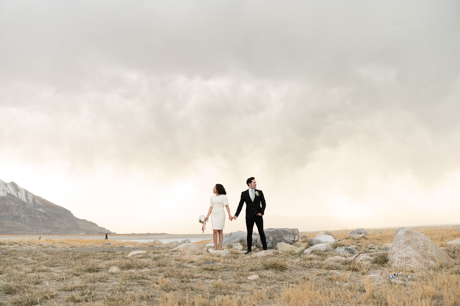 Couple holding hands in vast open field with dramatic cloudy sky and mountains in background.