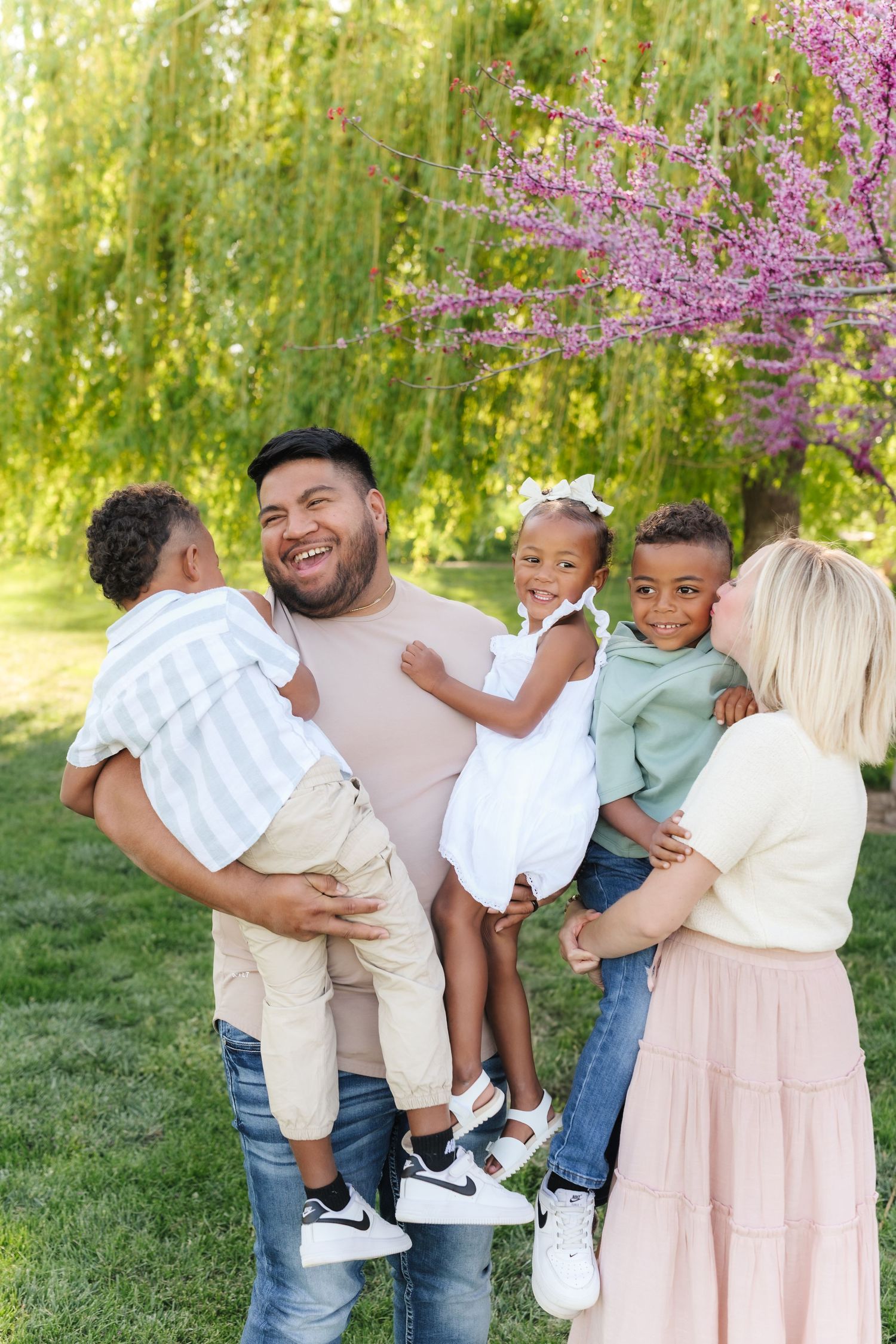 A family in coordinated pastel outfits embraces while standing in front of blooming cherry blossoms and willow trees.