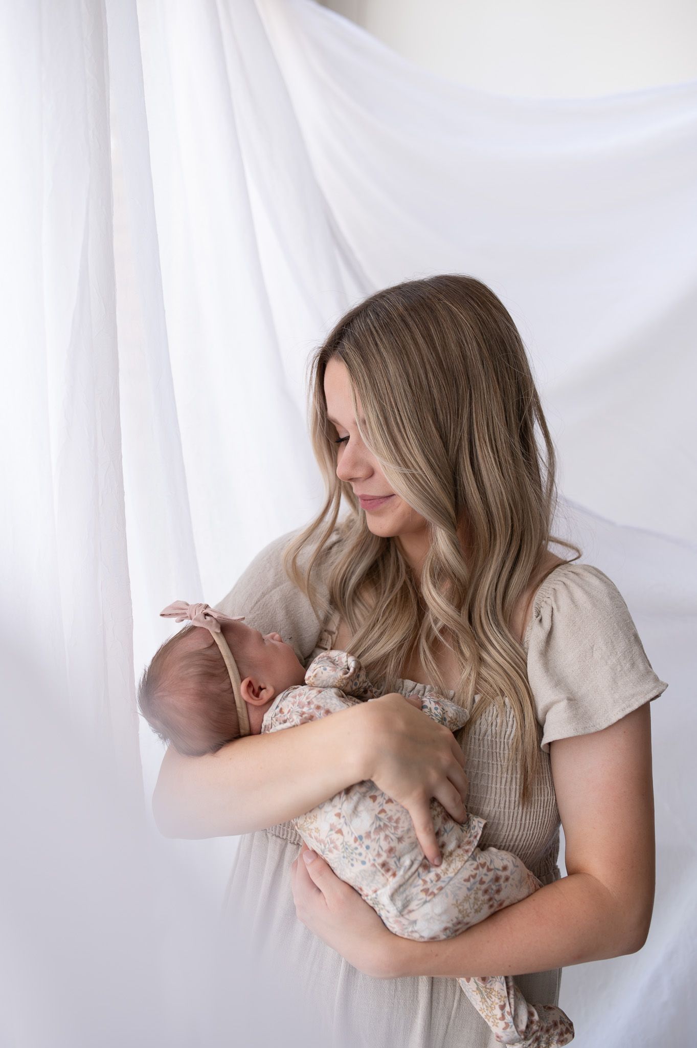 A mother tenderly holds her newborn baby in a bright, airy studio with soft white draping.