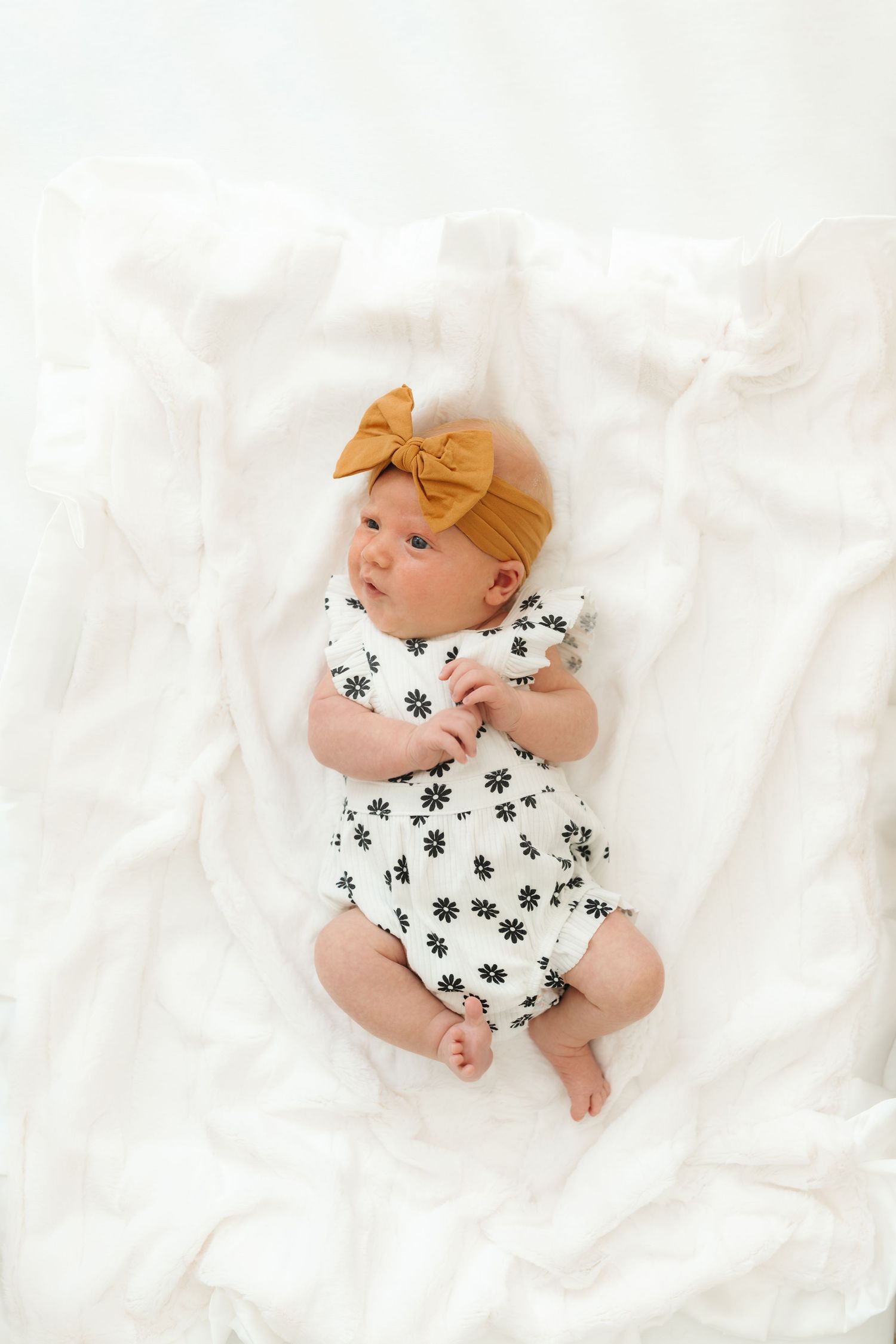 Baby wearing floral romper and mustard bow headband lying on white bedding.
