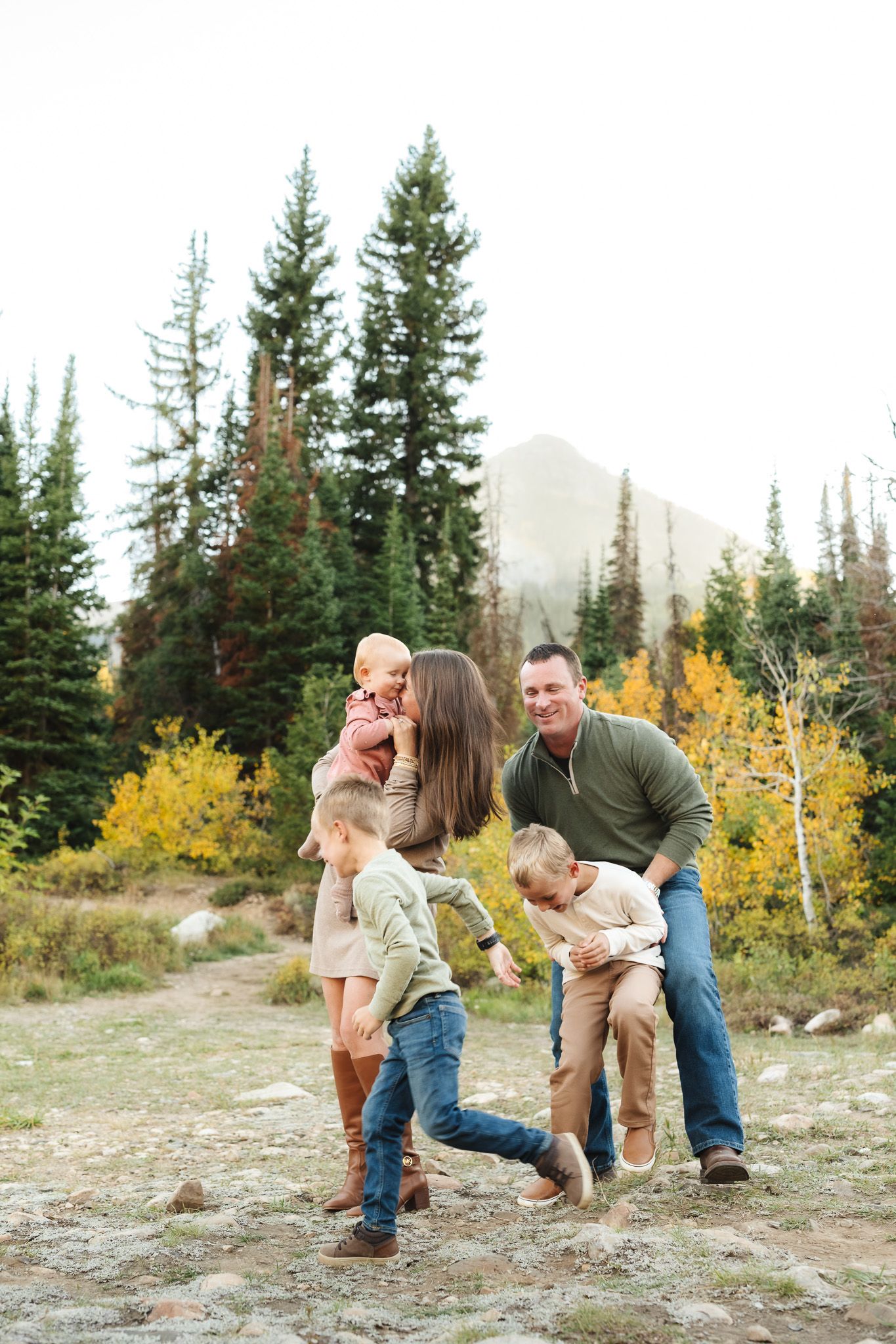 Family playing and laughing together in mountain setting with evergreen trees and fall colors.
