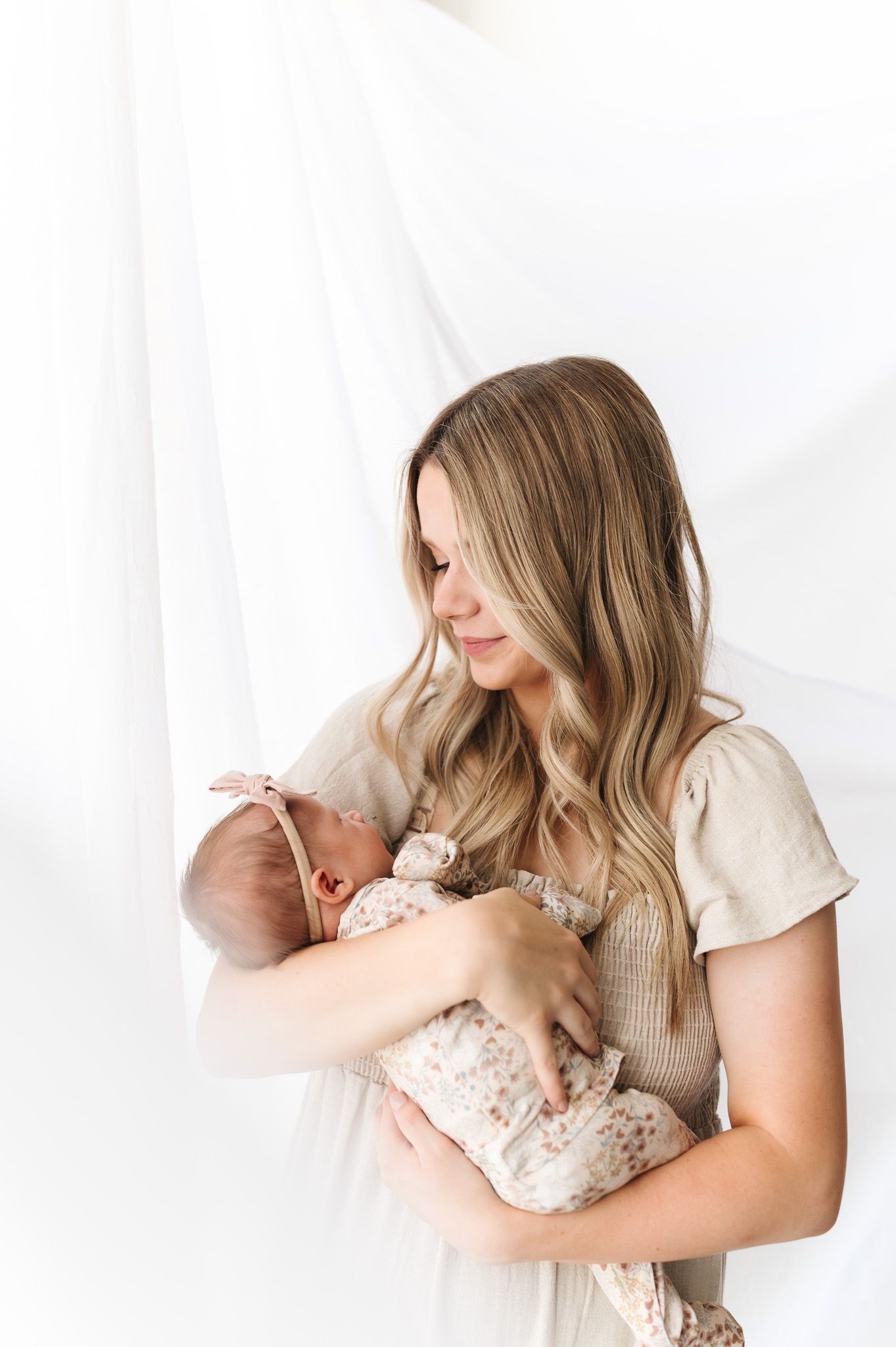 A mother in a beige dress tenderly holds her newborn baby against a soft white backdrop.