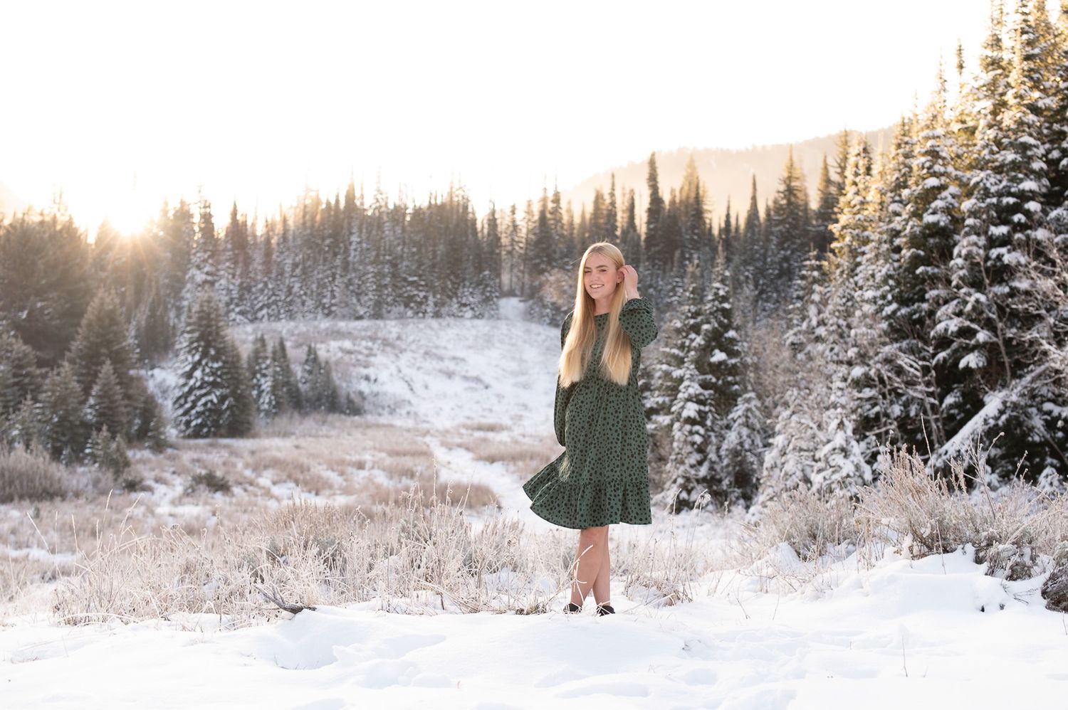 Girl in green dress standing in snowy mountain landscape with pine trees and golden sunset light.